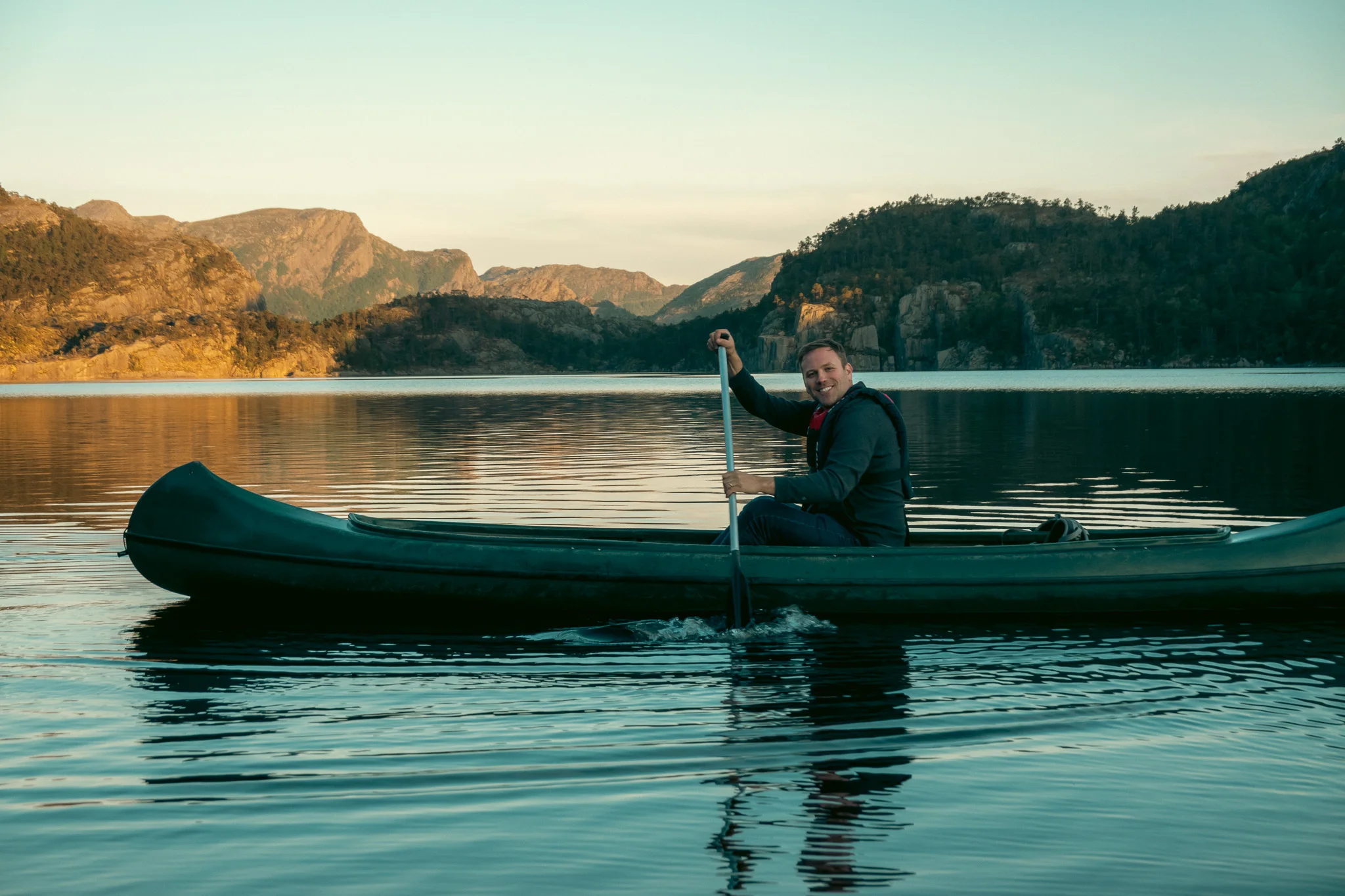 A man paddling a canoe on a lake with mountains in the background at sunset.
