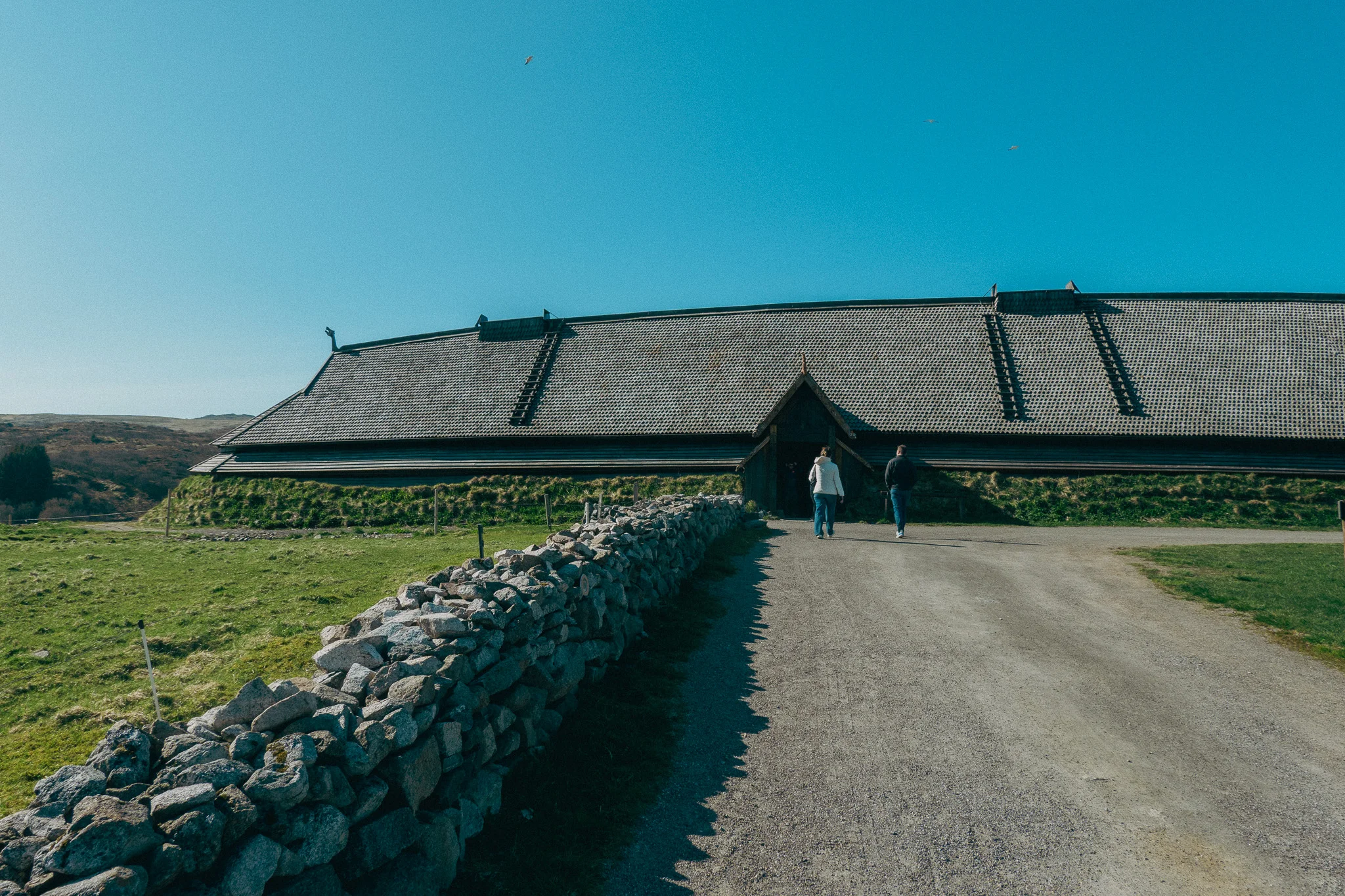 A gravel walkway lined with a stone wall leading to atraditional Viking Longhouse at the Lofotr Viking Museum