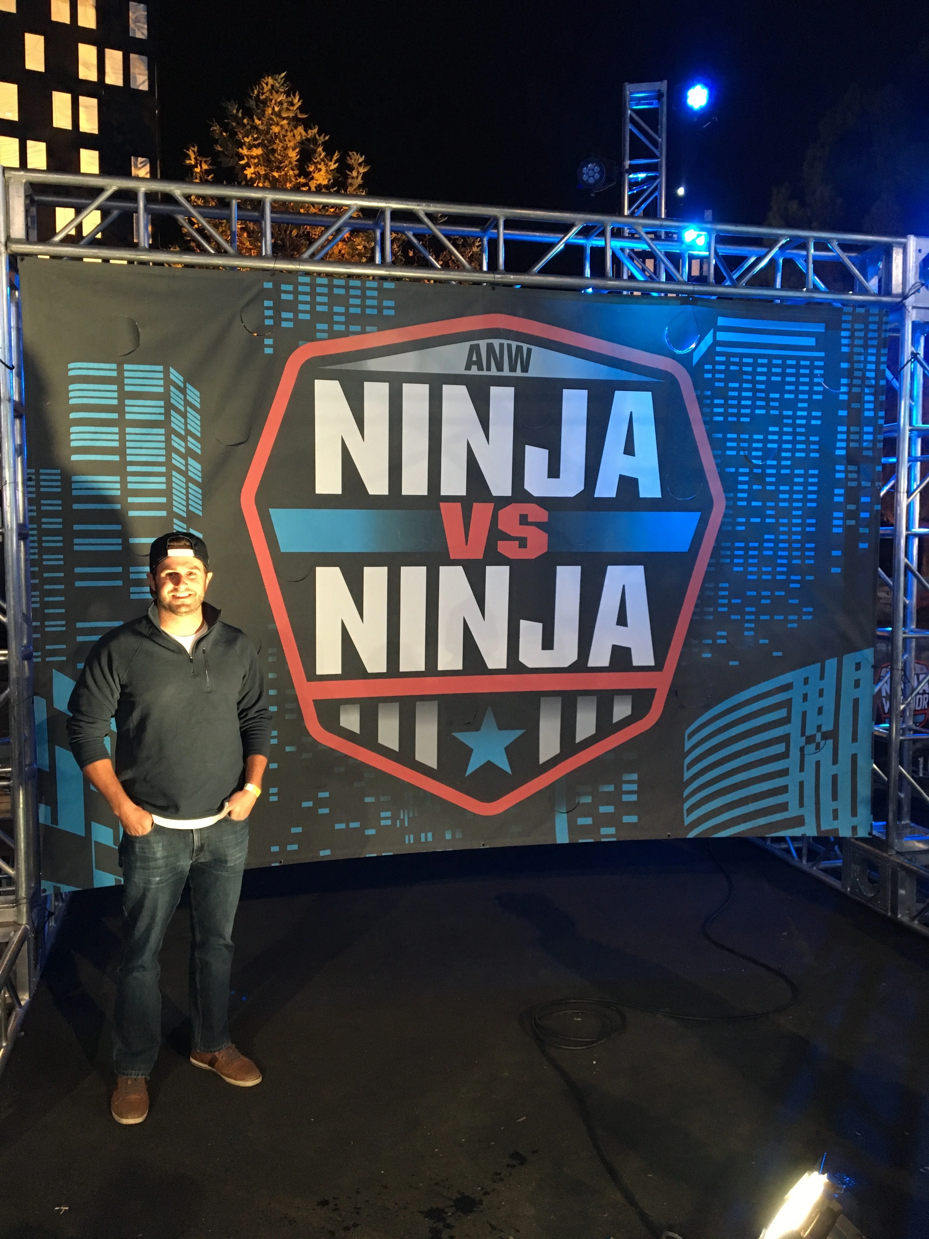Doug Newton posing in front of the American Ninja Warrior: Ninja vs Ninja logo he designed