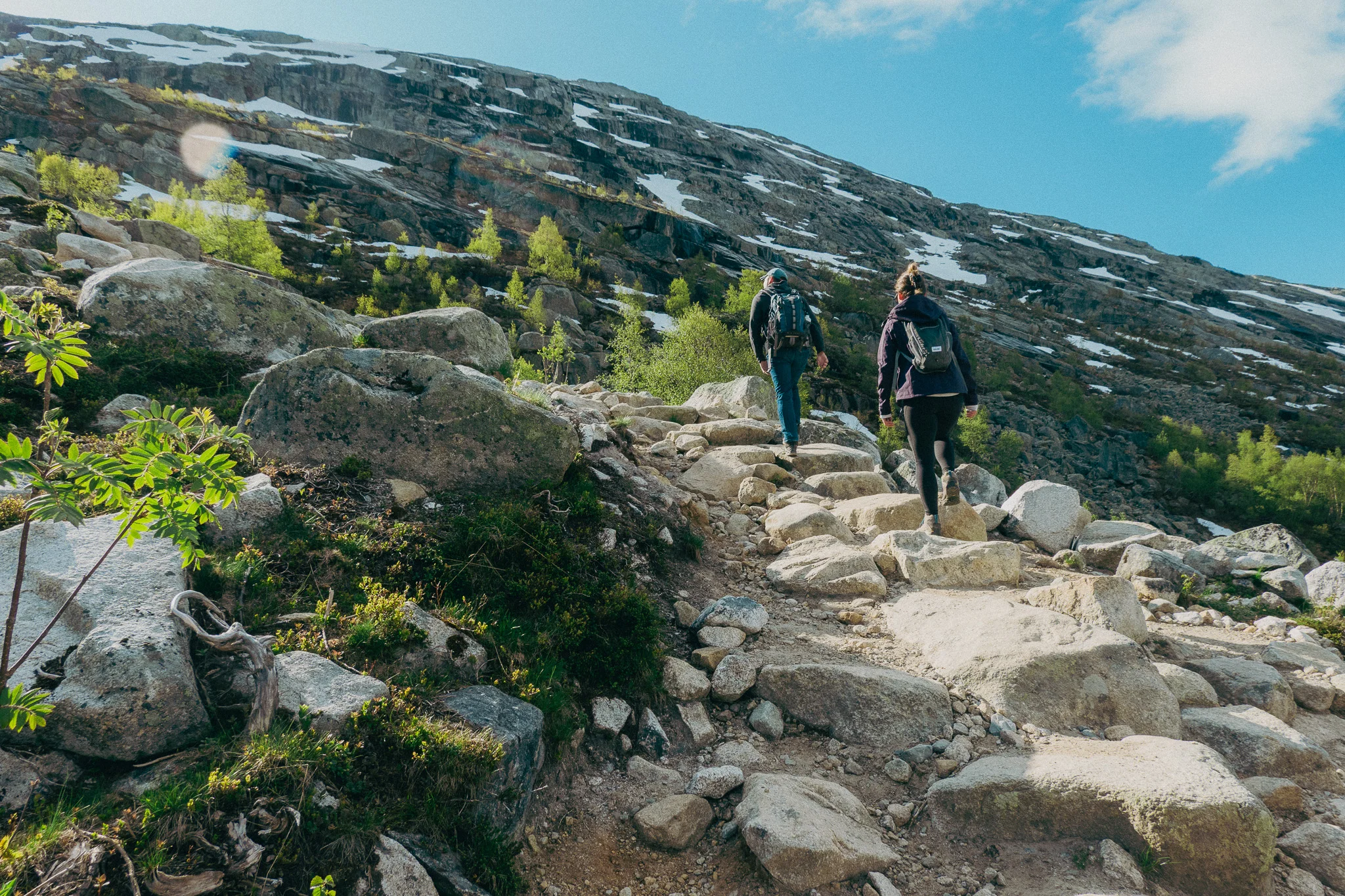 Two adults climbing a stone staircase on a hike in norway surround by rocks, green plants, and snow