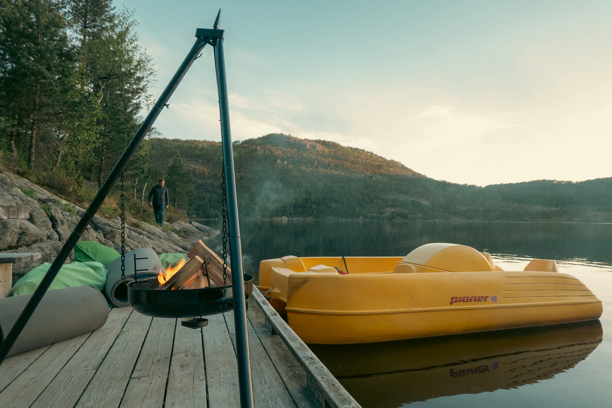 A fire pit on a lakeside dock and a boat floating on the water with trees and mountains in the background.
