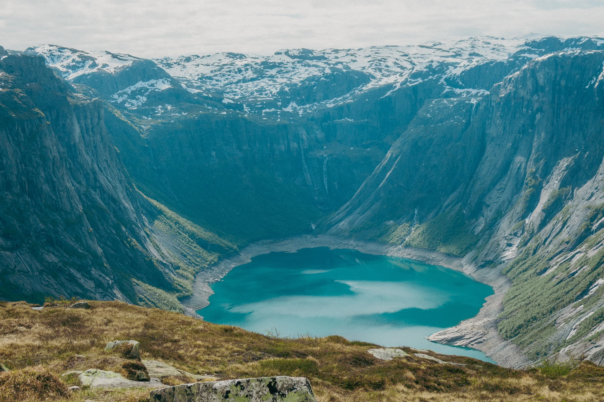 Scenic landscape with rocks and grass in the foreground and a blue lake and snowcapped mountains beneath a blue sky on the Trolltunga trail in Norway