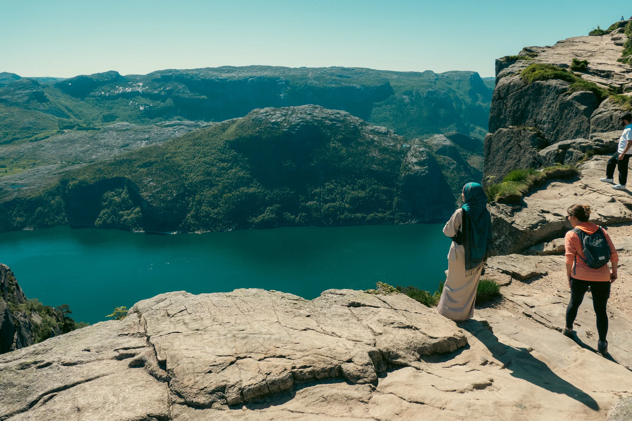 Two women looking over the edge of a cliff down at the fjord with mountains in the background