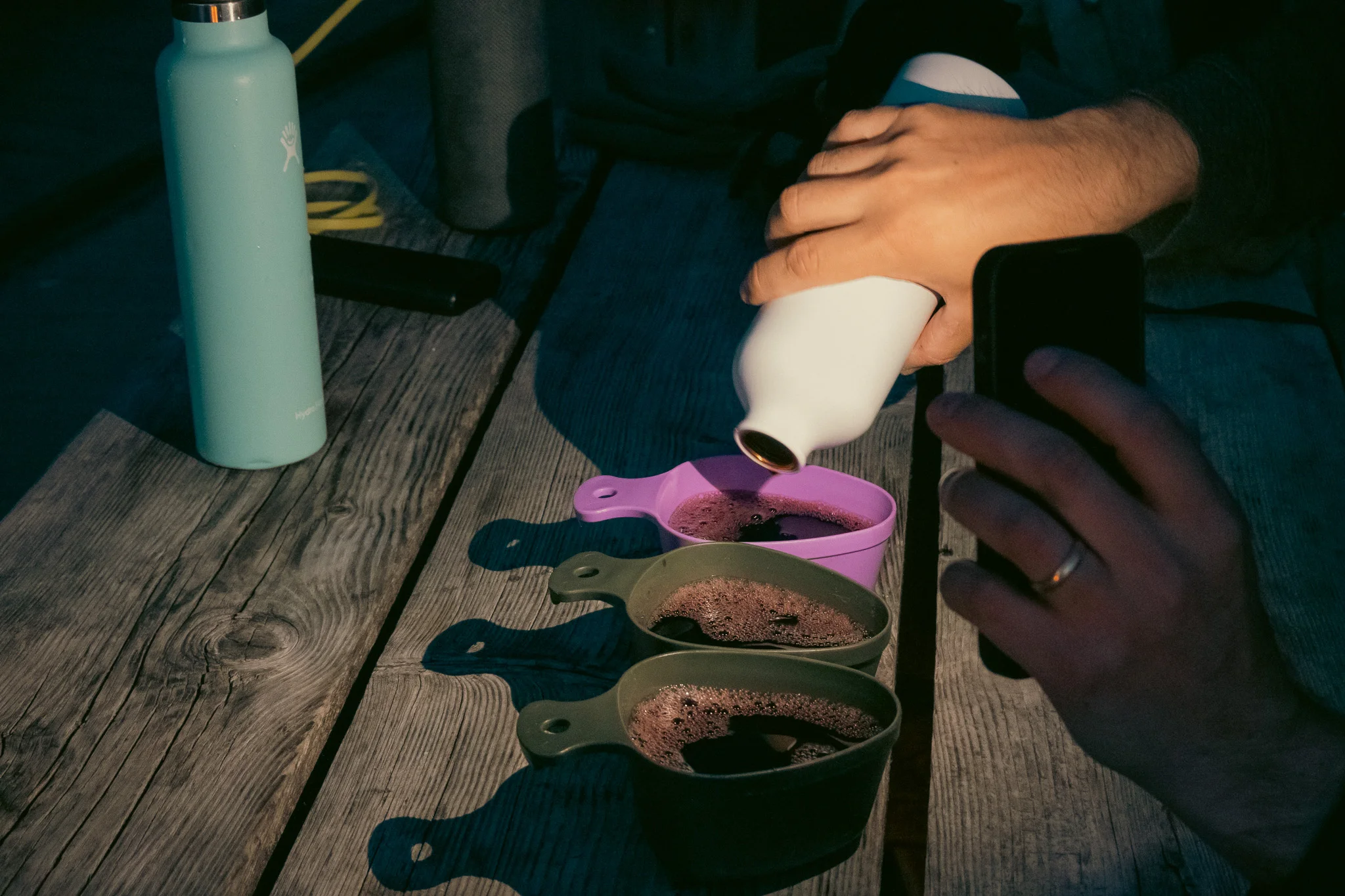 A hand pouring drinks from a water bottle into three large measuring cups at a picnic table at night.