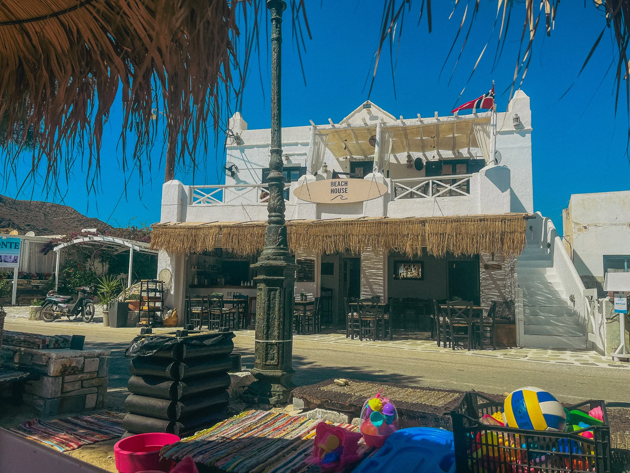 A white building with a sign that says Beach House with beach toys and straw umbrellas in the foreground