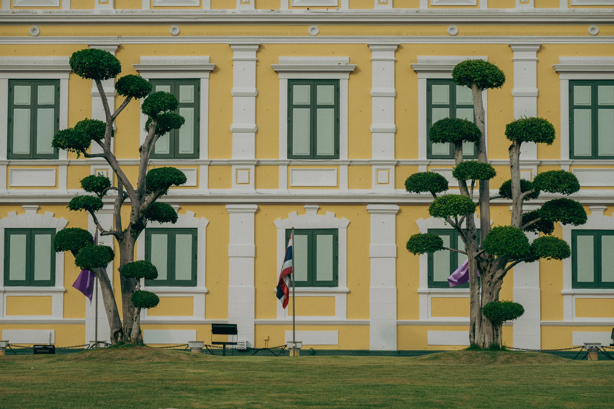 A bright yellow and white building with green trees and a Thai flag in the background