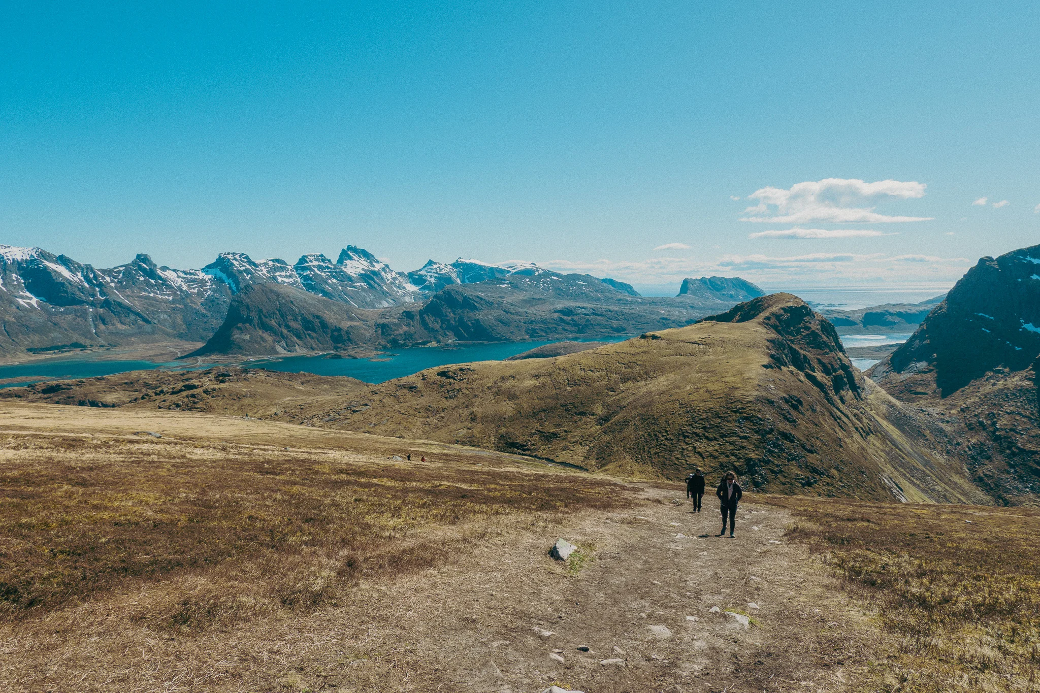 Two hikers stopped on a dirt trail with expansive snow-capped mountain views and blue sky in the background