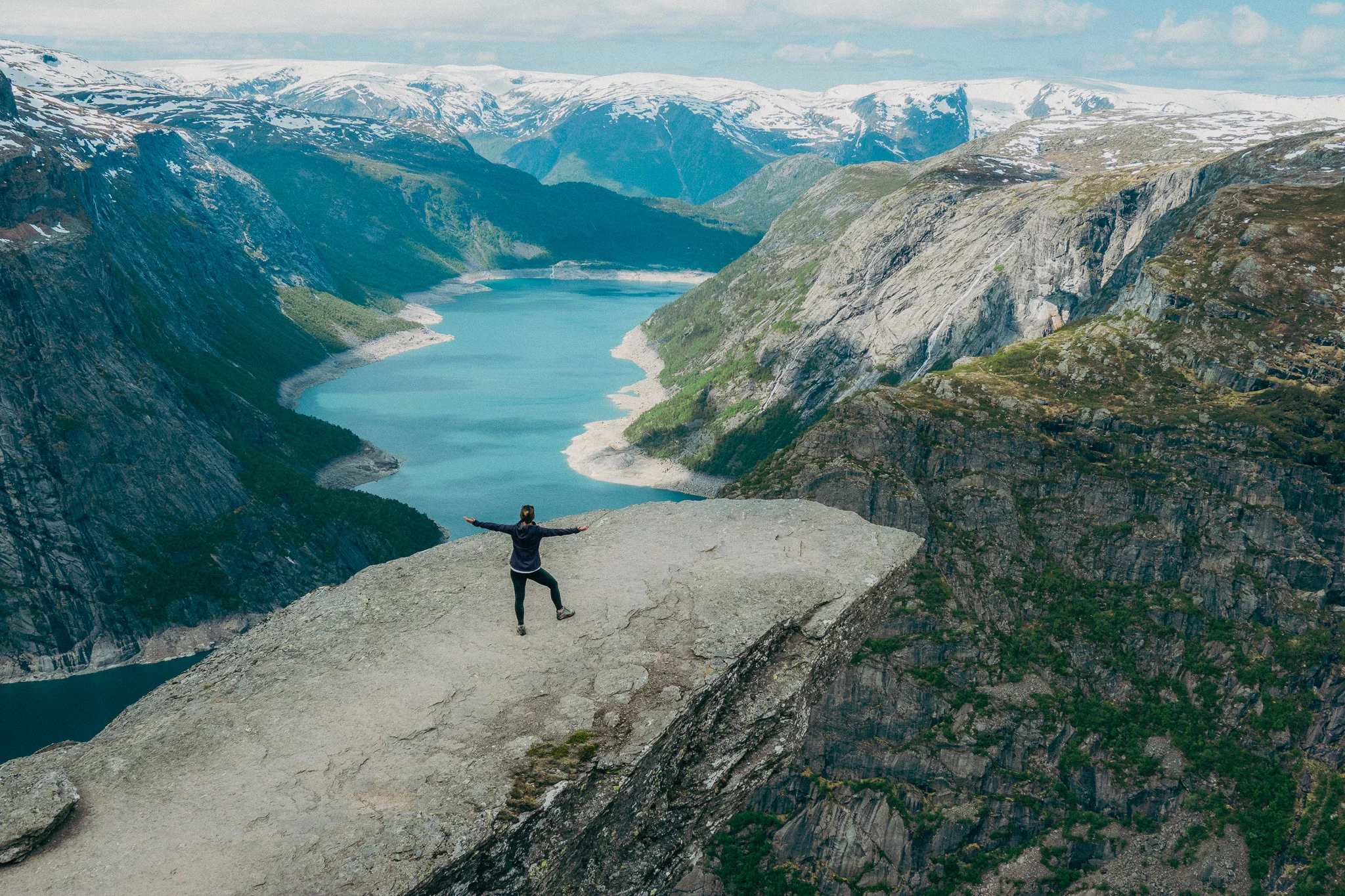Adult female stands at the tip of Trolltunga in Norway with snowcapped mountains and blue water in the background