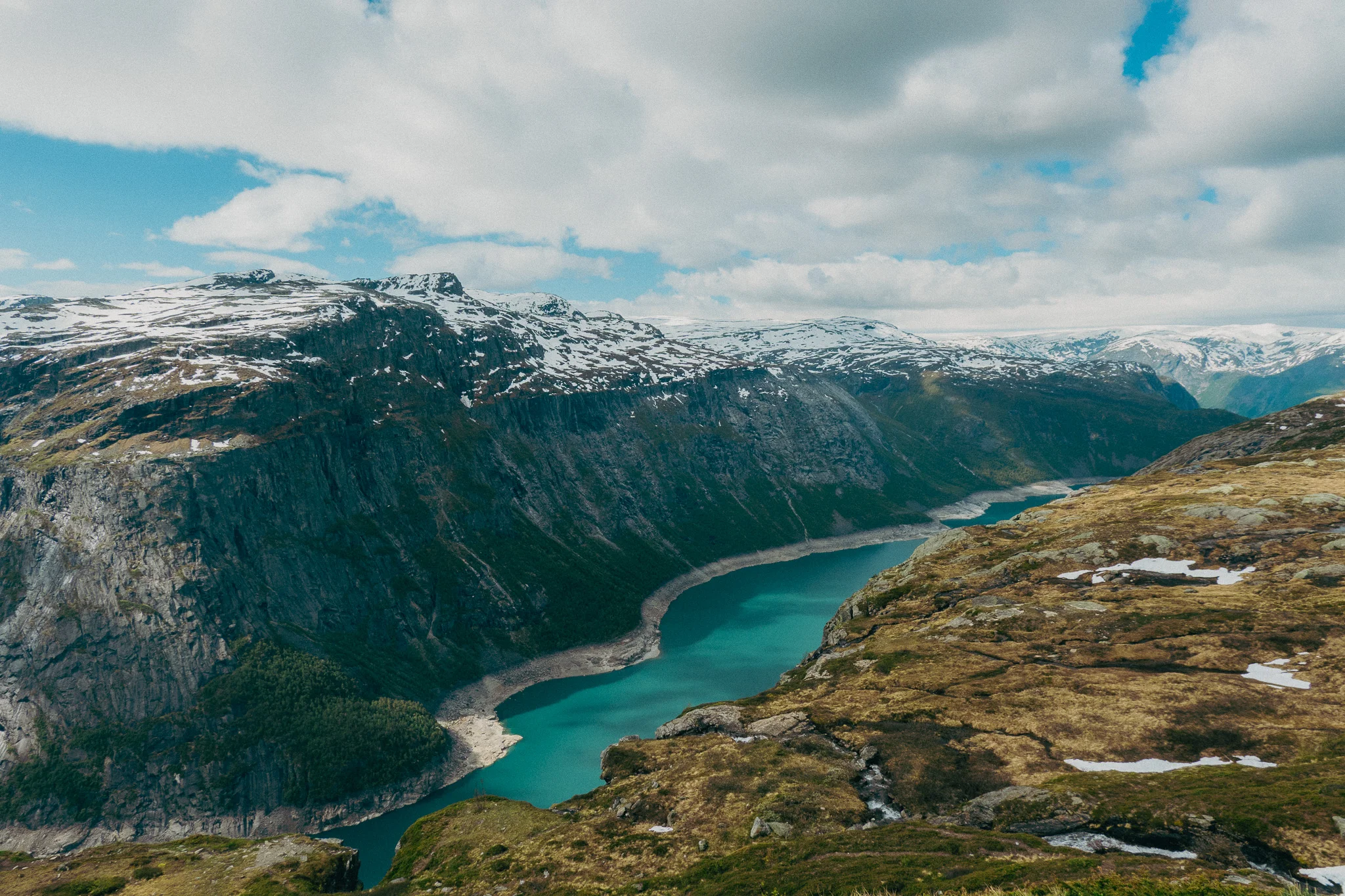 Scenic landscape with rocks and grass in the foreground and a blue lake and snowcapped mountains beneath a blue sky on the Trolltunga trail in Norway