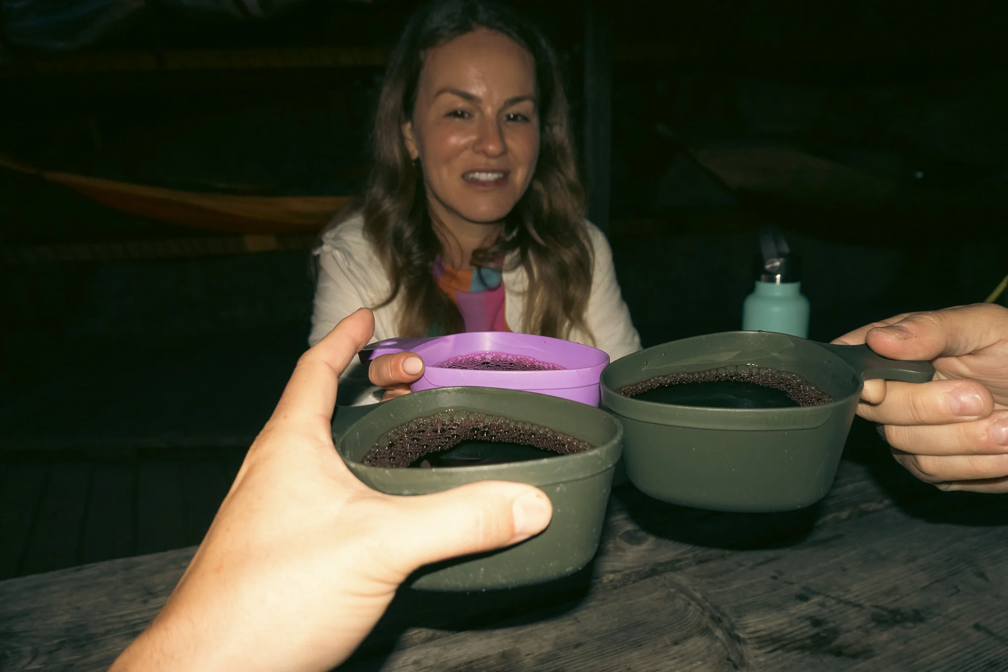 Three people toasting drinks in large measuring cups at a picnic table at night.