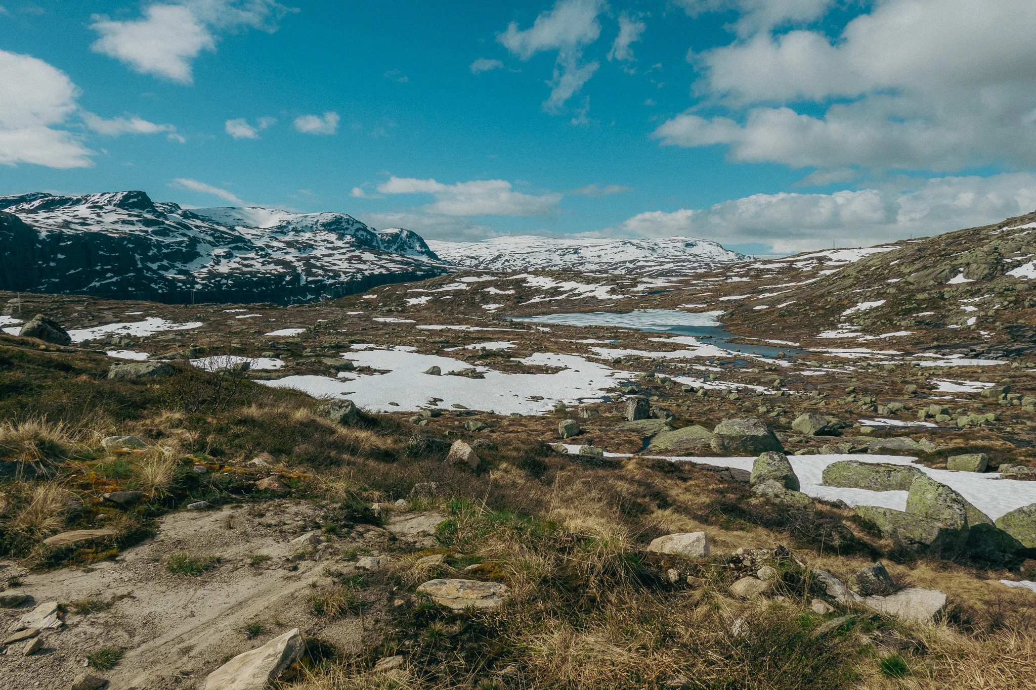 Scenic landscape with rocks, brush and snow in front of snowcapped mountains and blue skies on the Trolltunga trail in Norway