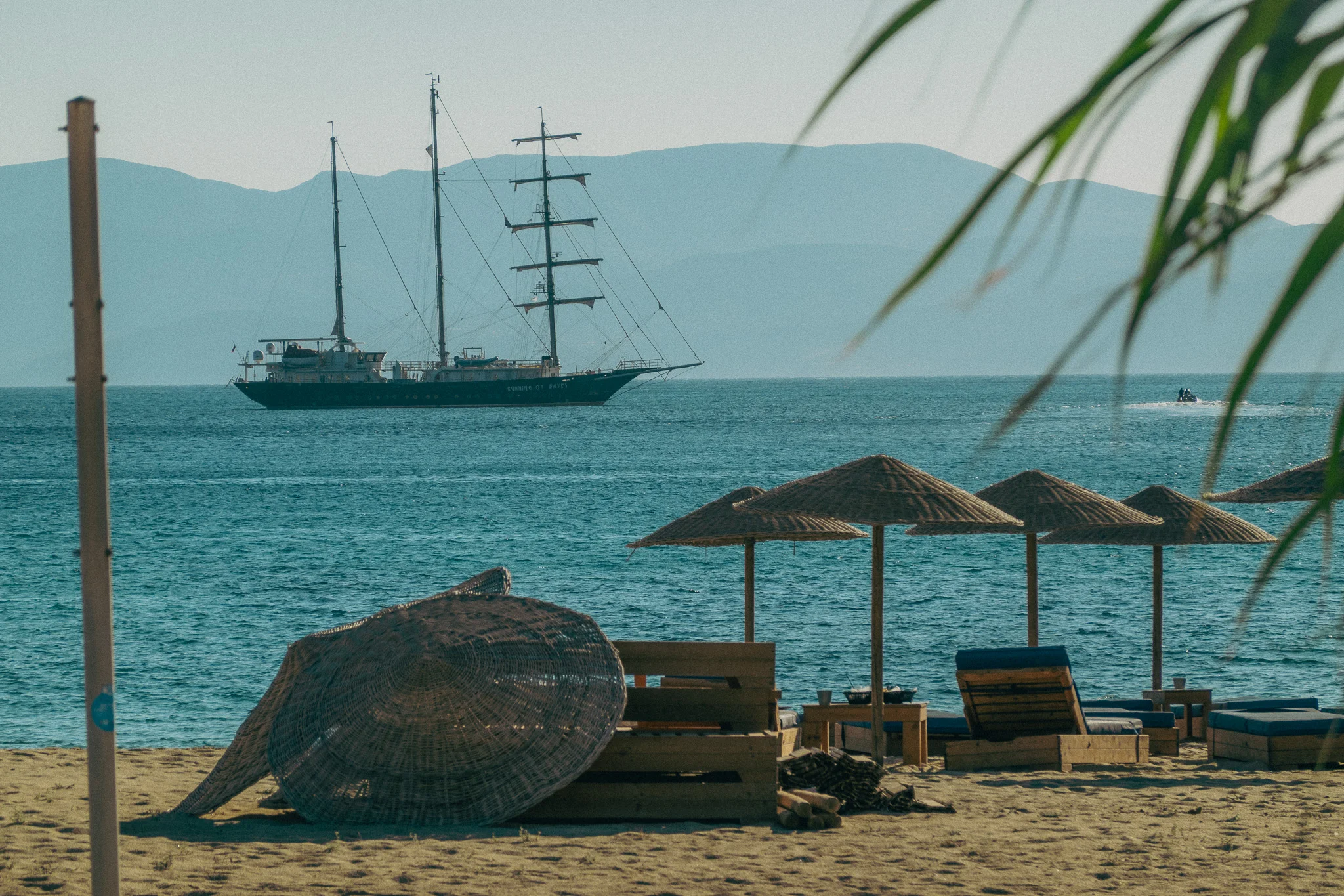 Beach chairs and straw umbrellas on the beach with a palm tree in the foreground and a tall sailing ship on the water in the background