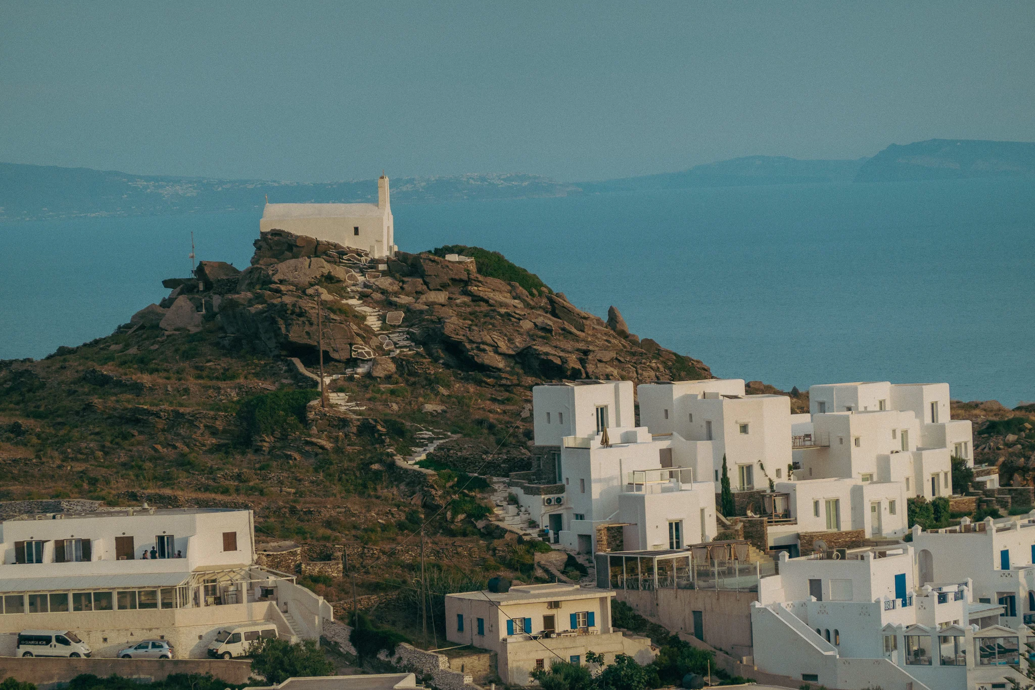 A small chapel perched atop a hill surrounded by white building and ocean in the background