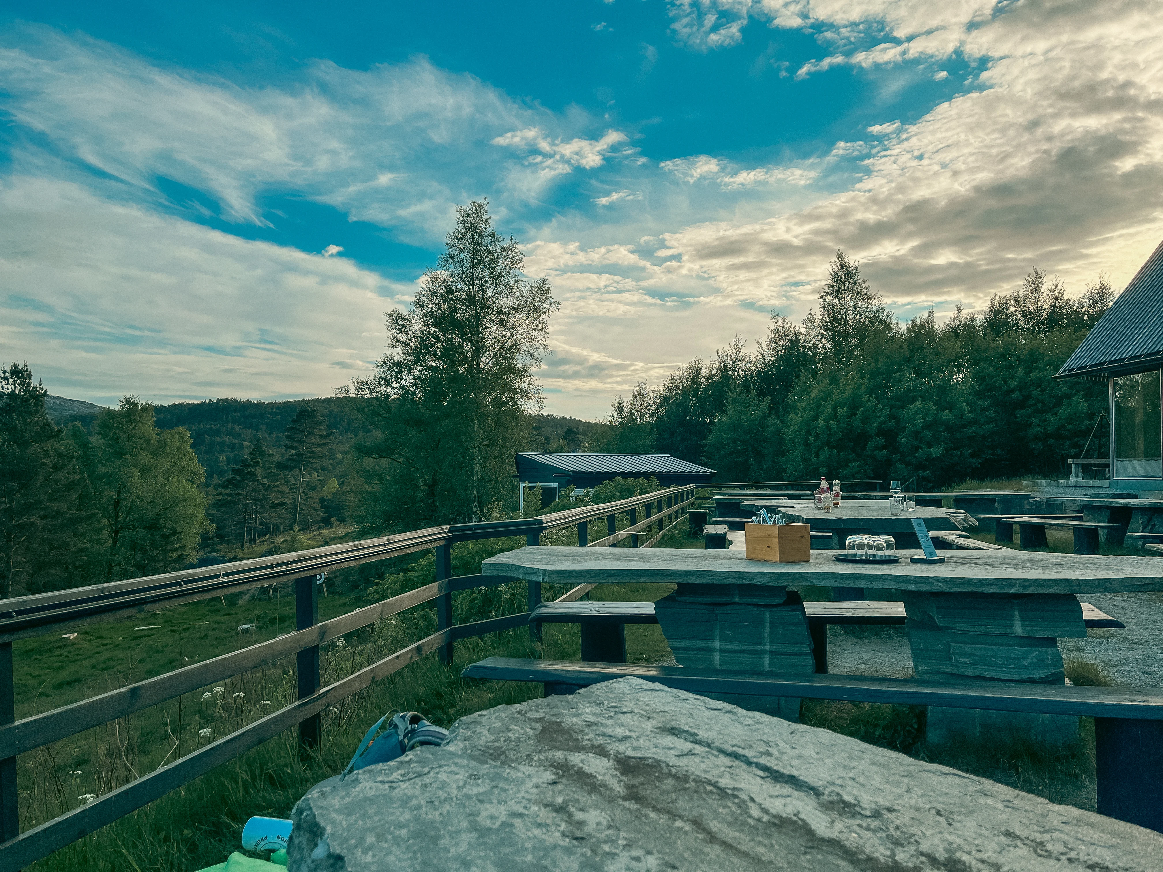 Stone picnic tables on a dining patio lined by a fence with trees and mountains in the background.