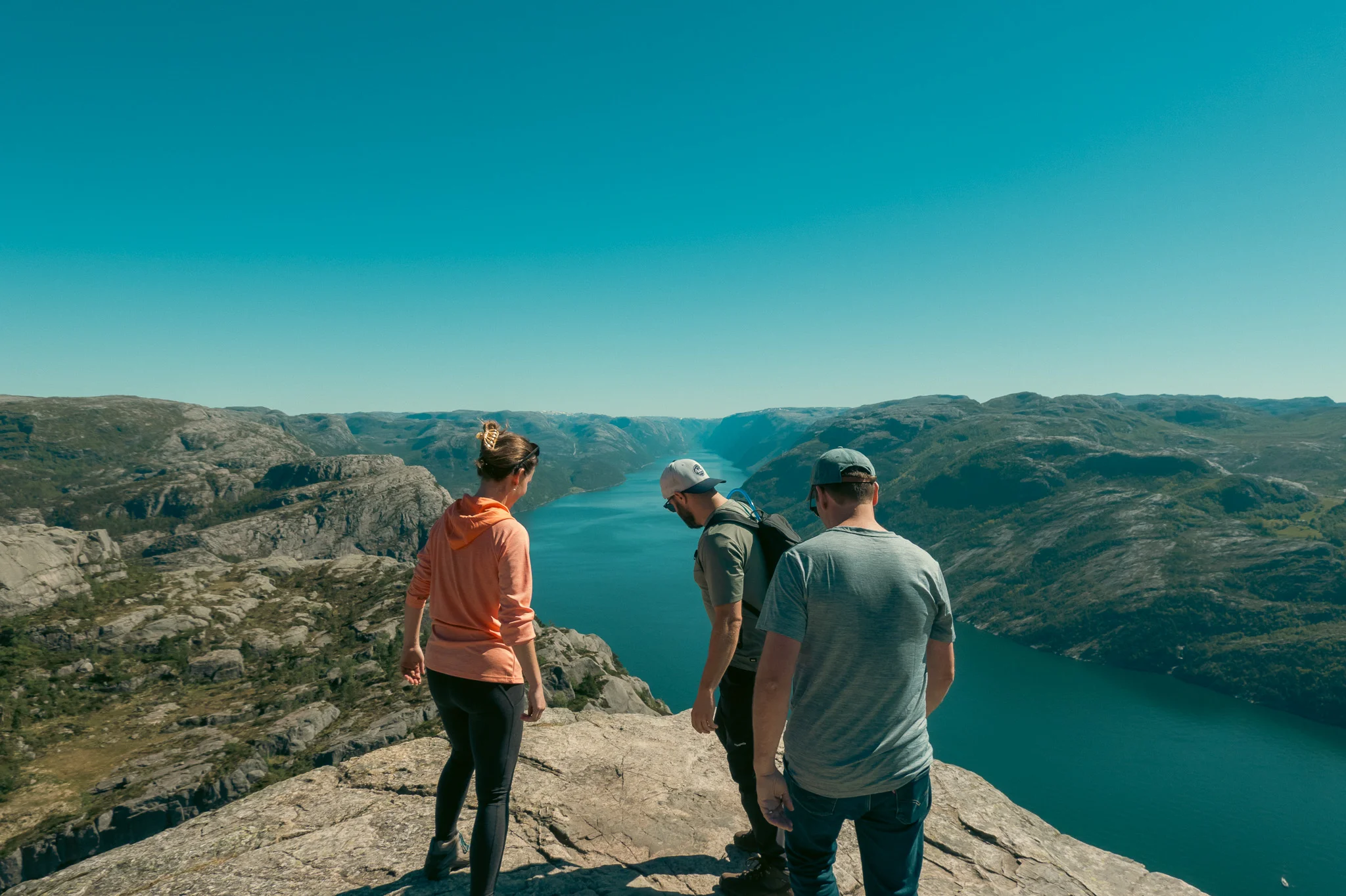 Two men and a woman approach the ledge at Pulpit Rock with the fjord and mountains in the background