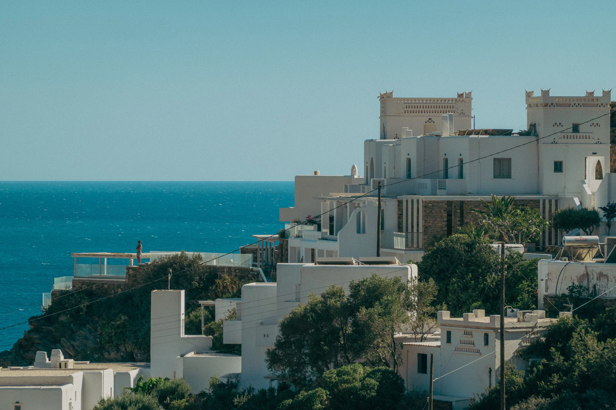 White buildings on the side of the mountain overlooking the ocean with a man standing on the balcony looking off into the distance.