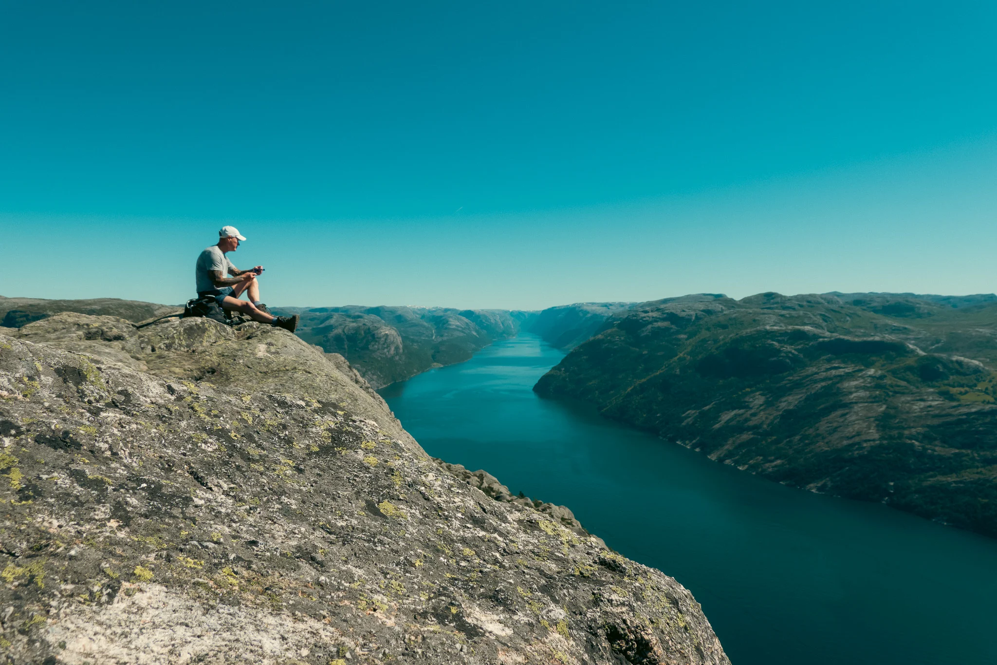 A man sits on top of a cliff with the fjord and mountains in the background in Norway.