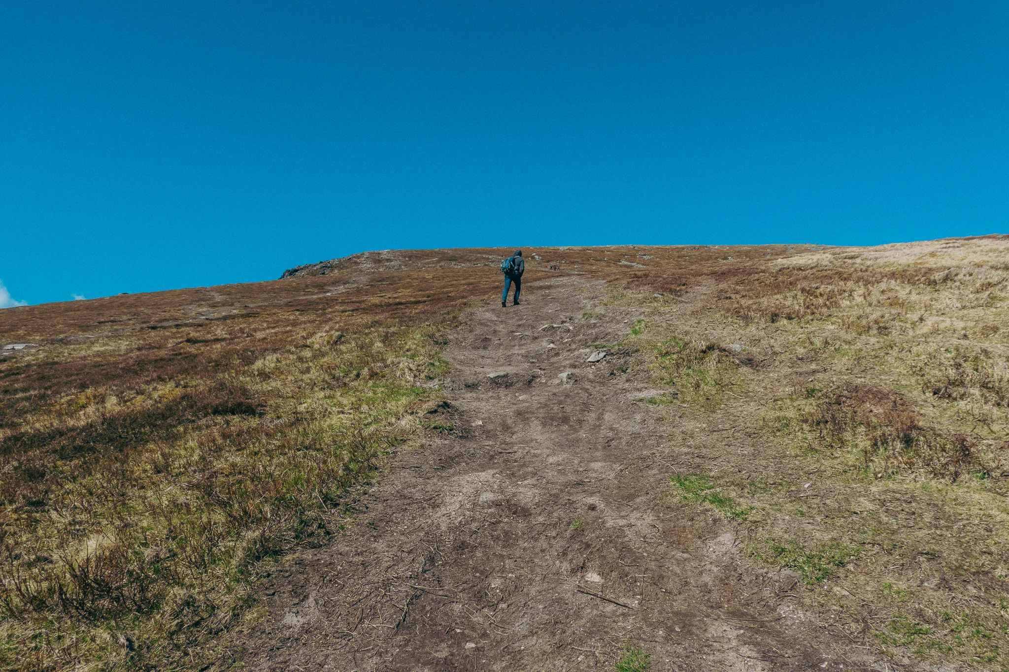 A hiker climbs a steep uphill dirt trail surrounded by grass and blue sky