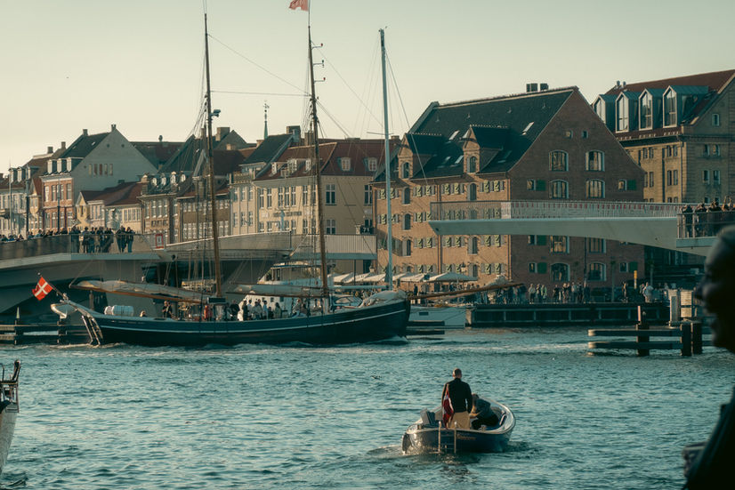 A small wooden boat leaves the doc on a river with a large sail boat crossing through a draw bridge in the background in Copenhagen, Denmark