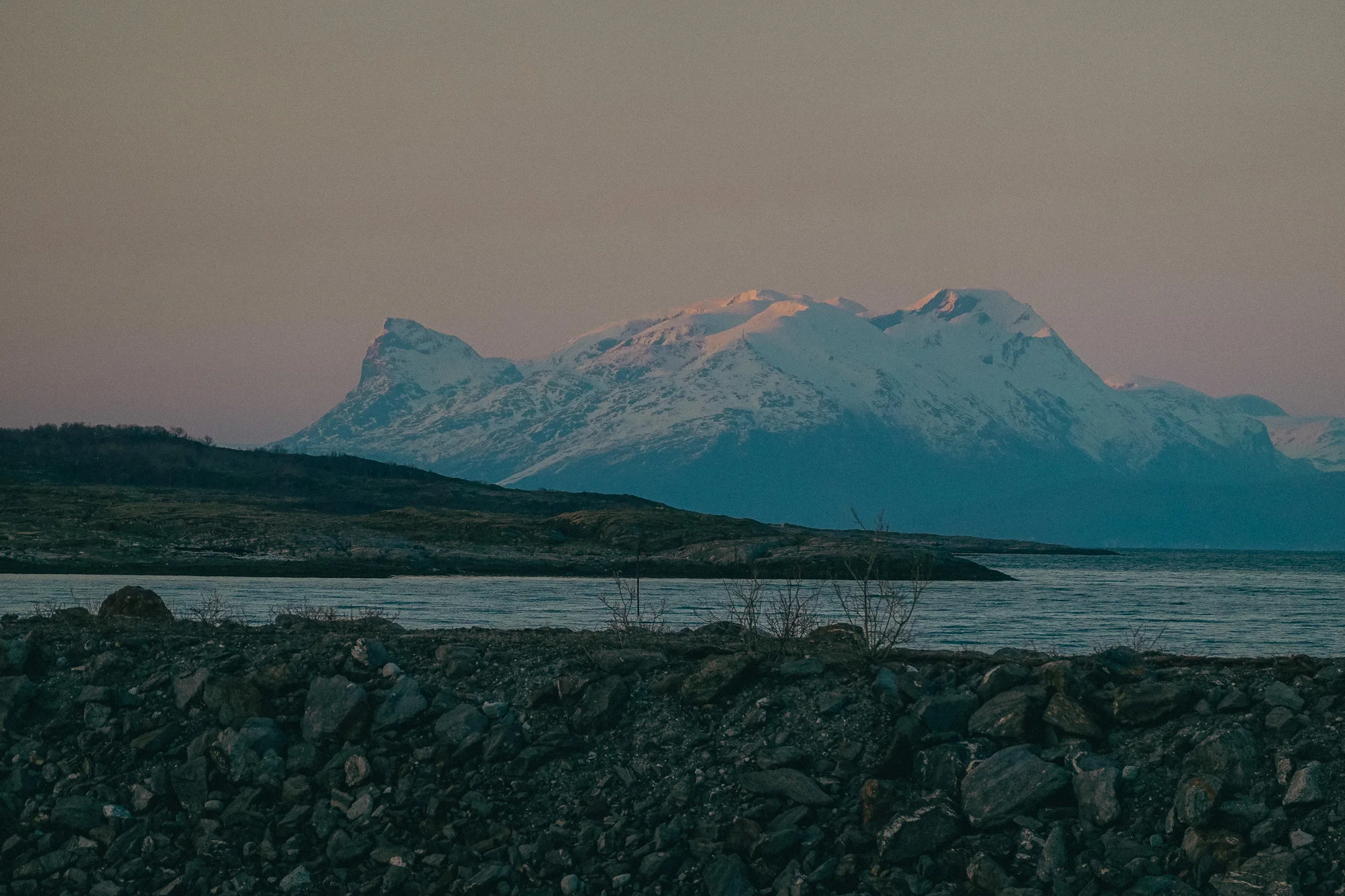 Snow-capped mountain landscape with water and rocky land in the foreground at dusk in Narvick Norway