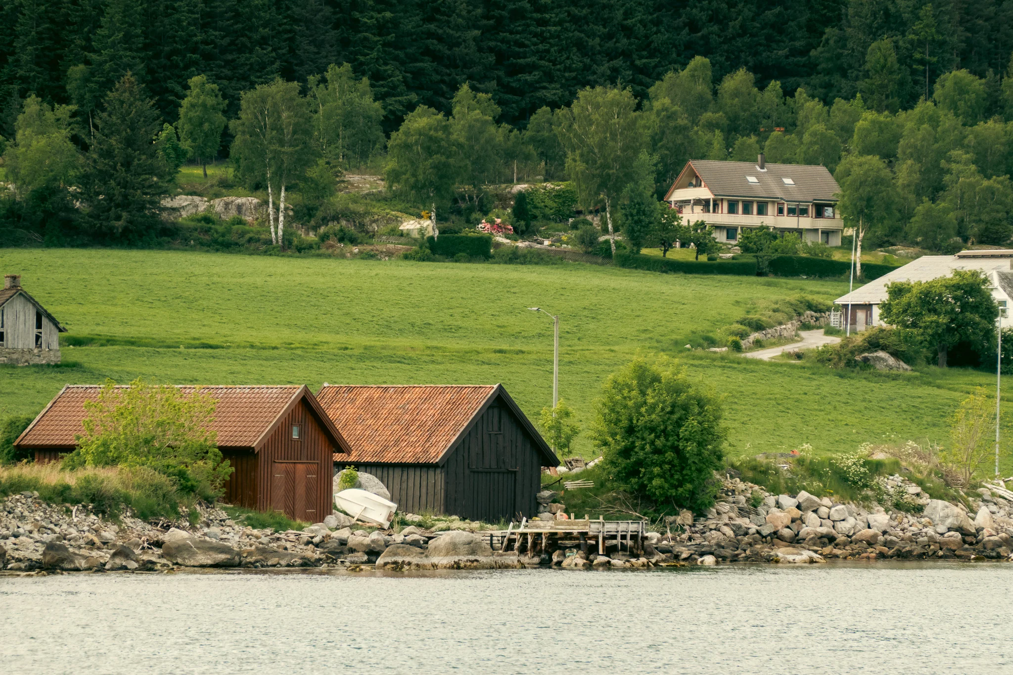 Two barns on the shore of a fjord with bright green grass and a house in the background.