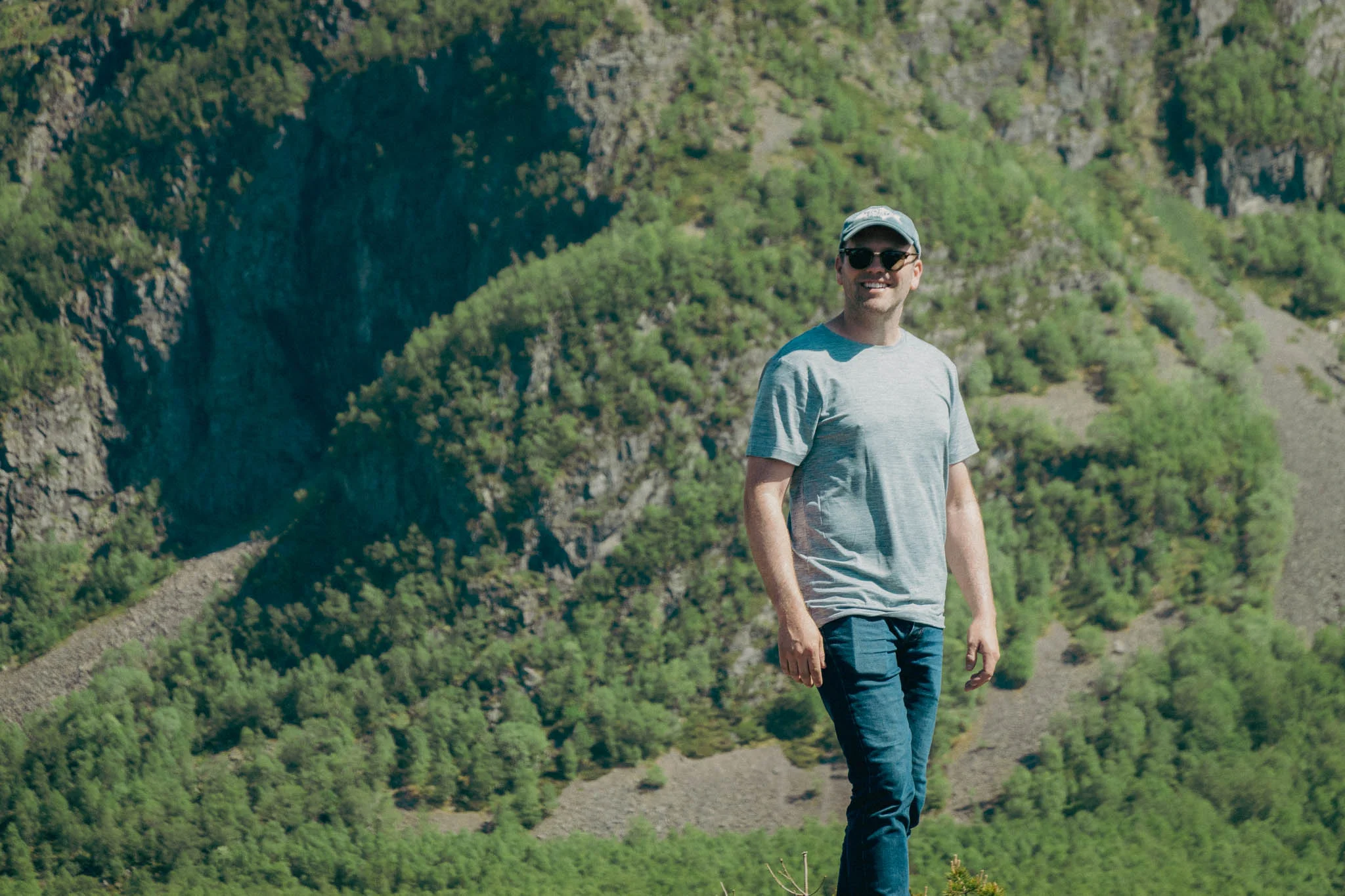 Man standing on a rock in front of green mountains wearing a hat and sunglasses