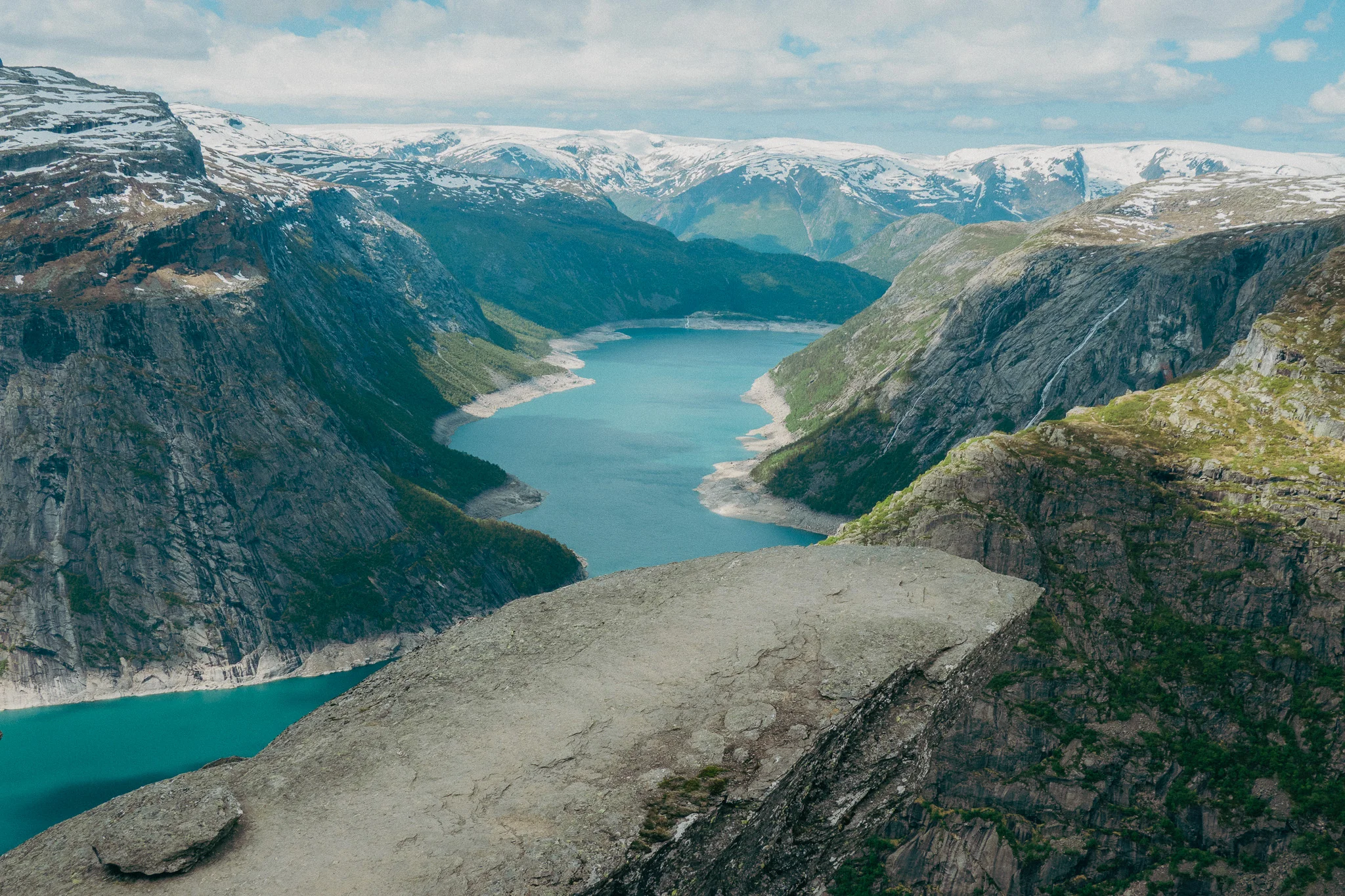 Trolltunga rock in Norway with blue water, snowcapped mountains, and partly cloudy skies in the background