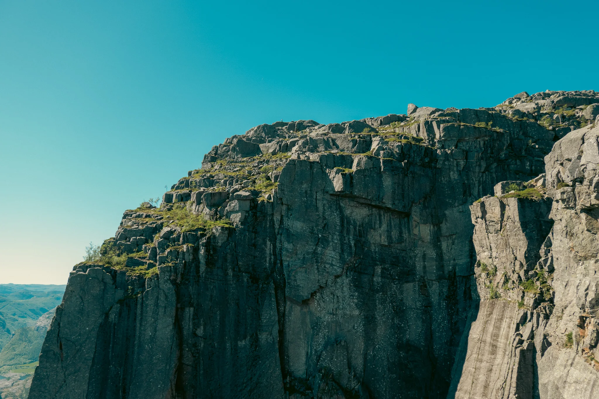 A jagged cliff lined with trees with blue sky and mountains in the background in Norway.