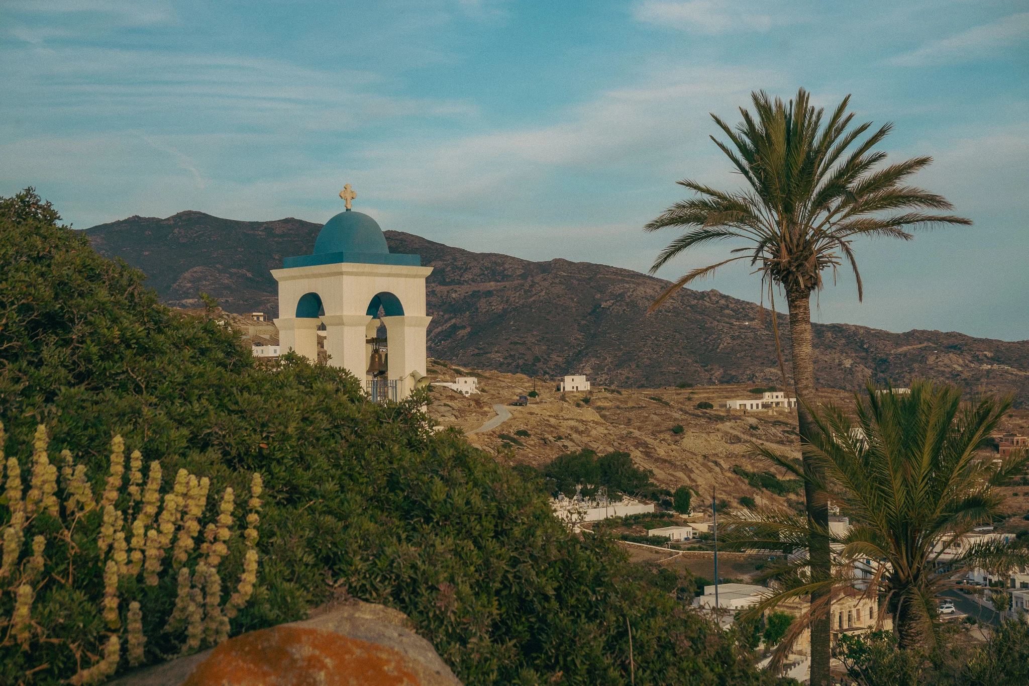 A white and blue chapel with mountains and blue sky in the background and a palm tree and vegetation in the foreground