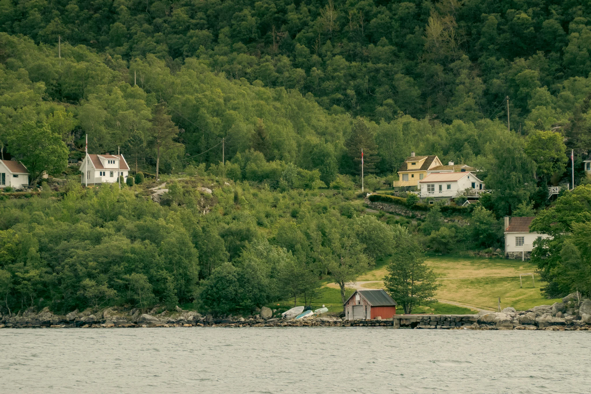 A red shed and white buildings alongside a fjord surrounded by green trees.