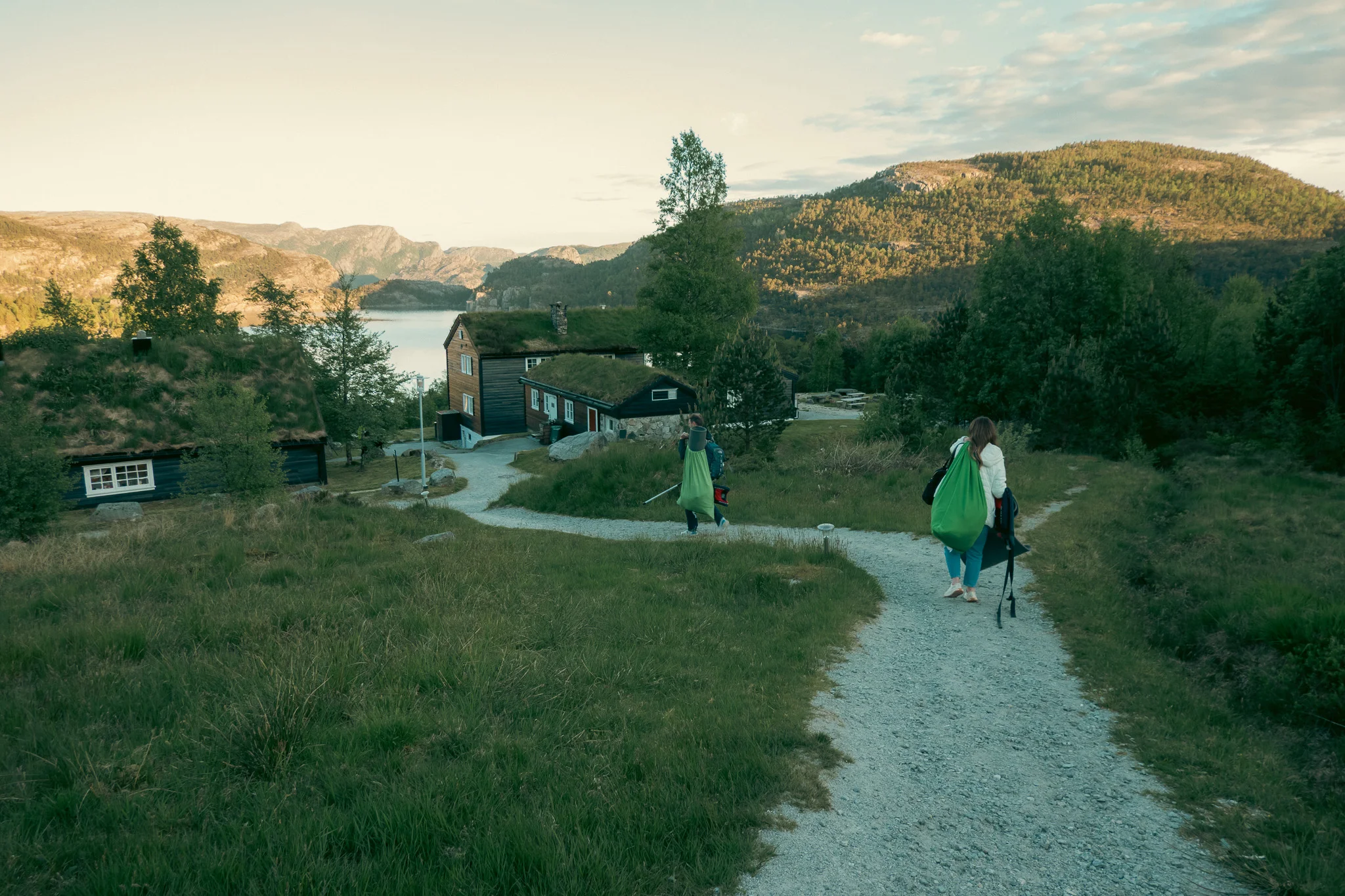 A man and woman holding camping gear and a canoe padle walking down a path towards a lake with mountains in the background at sunset.