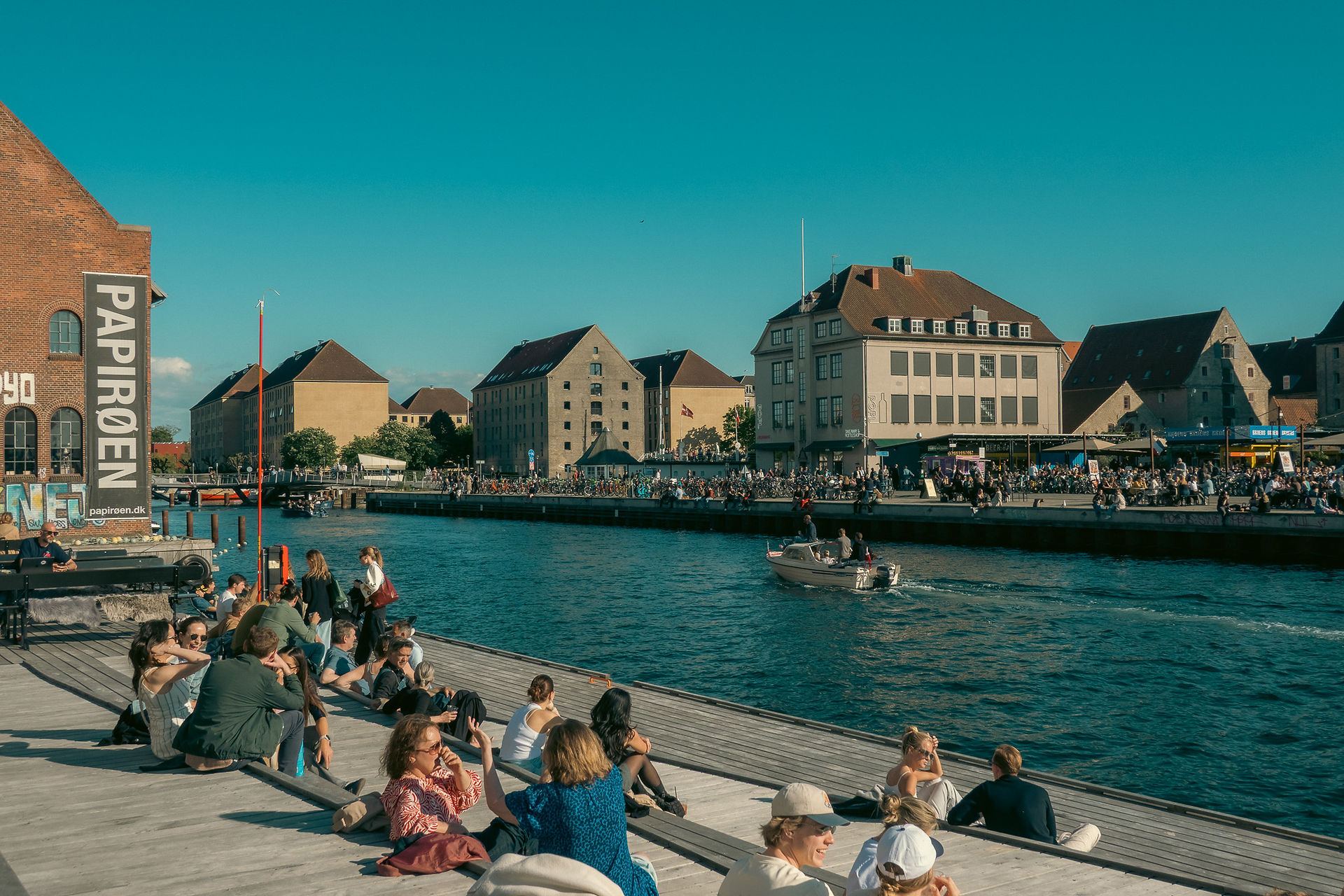 Several groups of people sitting on a wooden lounge area along the river with a sailboat going by in Copenhagen, Denmark