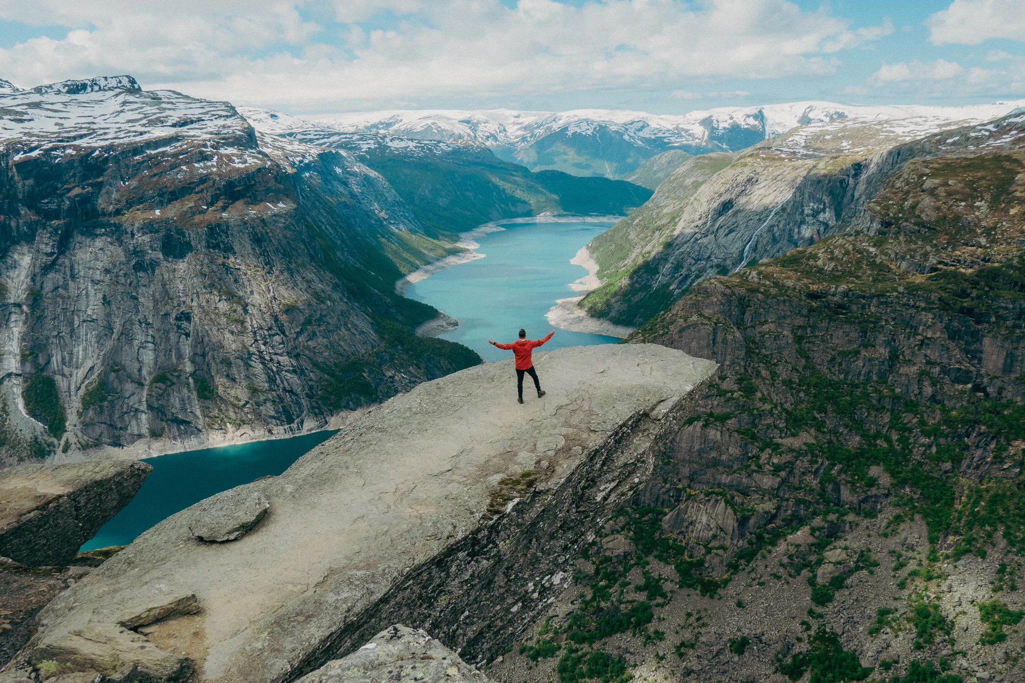 Adult male stands at the tip of Trolltunga in Norway with snowcapped mountains and blue water in the background