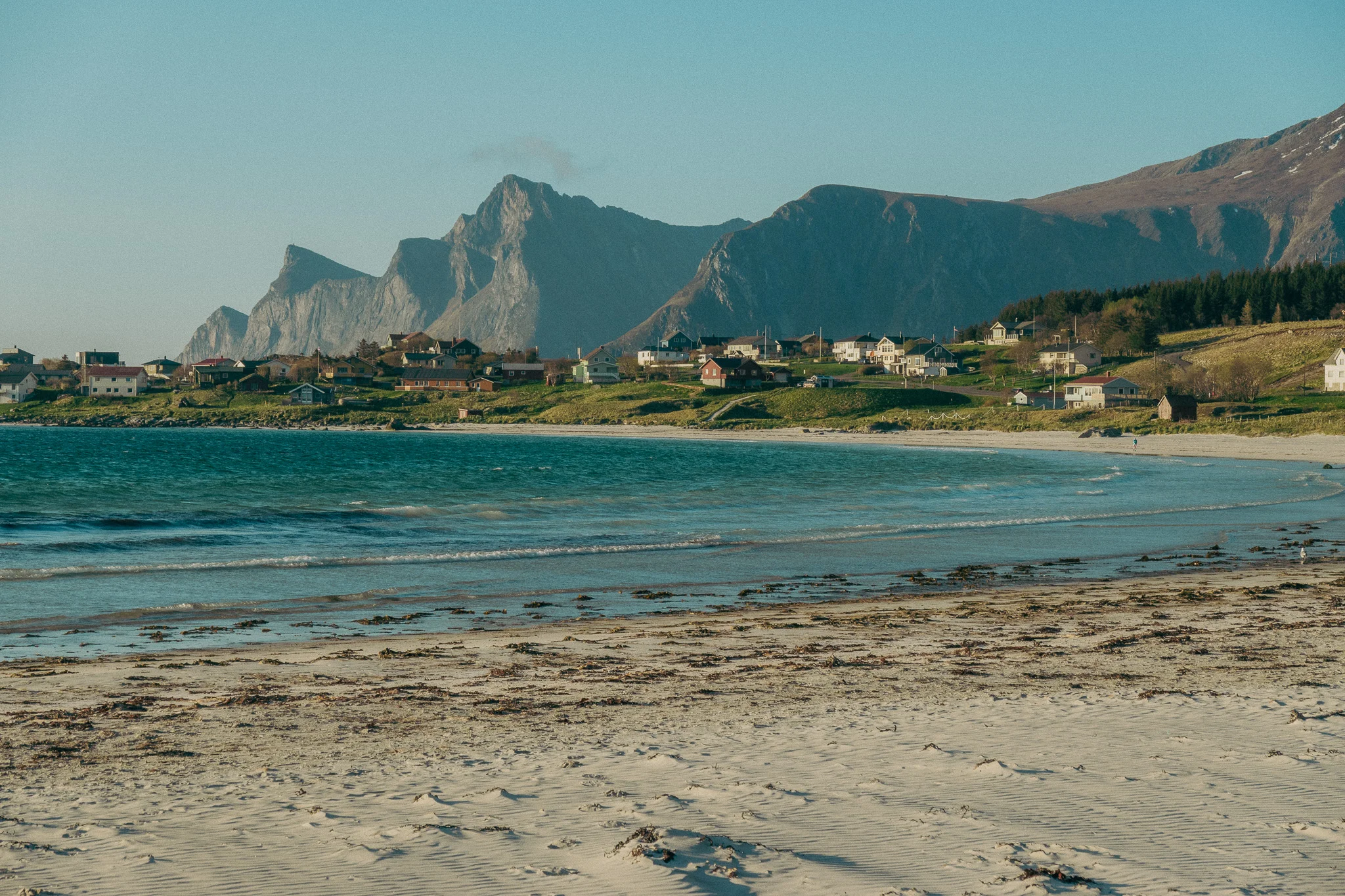 White sand beach shore with cottages on a grassy hillside along the water and jagged mountains in the background