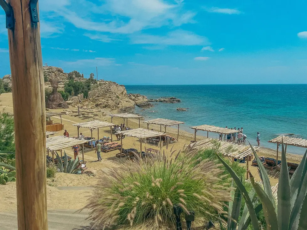 Beach scene with people under wooden canopies on sandy shore. Blue sea and sky, rocky cliffs in the background. Relaxed, sunny atmosphere.