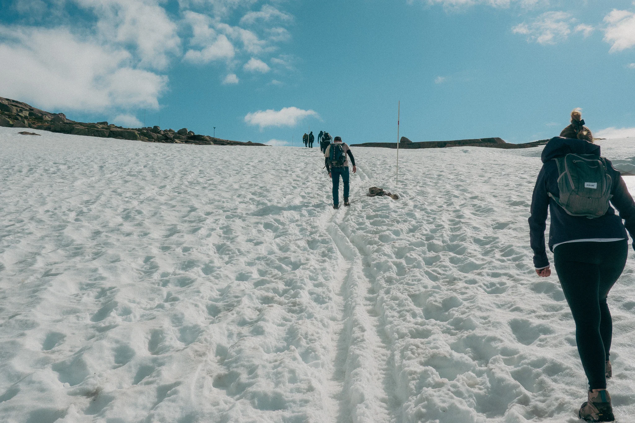 A line of adults hiking through snow surrounded by rocks and blue sky in the background on the Trolltunga trail in Norway