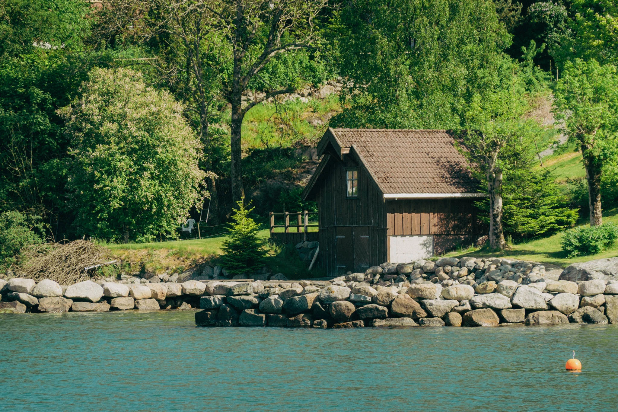 Small brown barn along the water surrounded by green sunlit trees