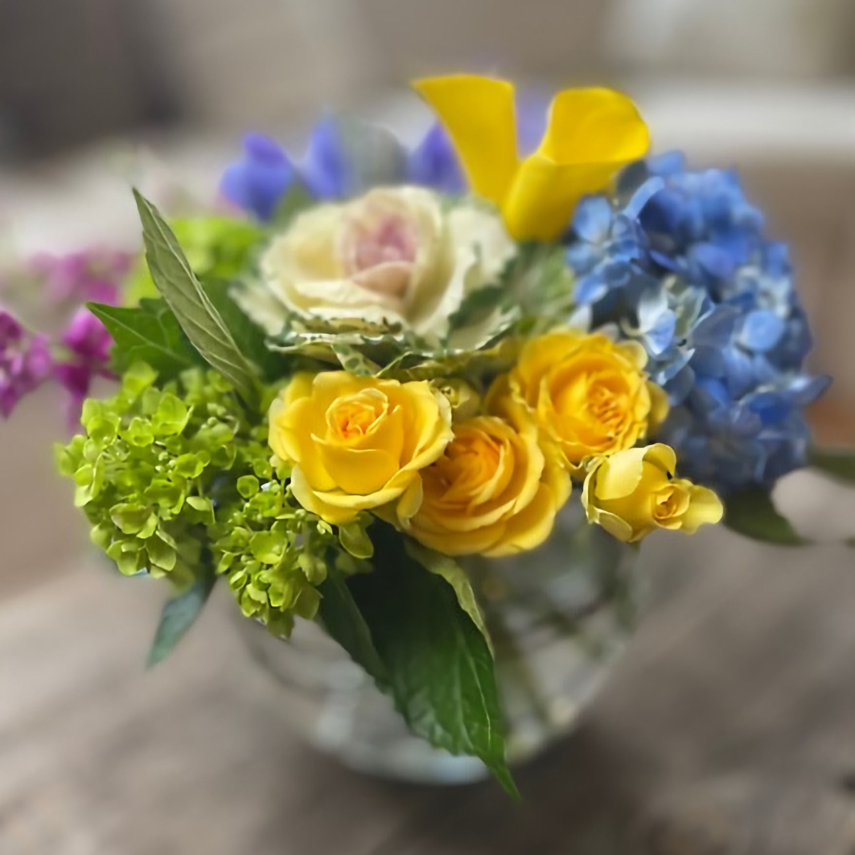 8 inch round bubble bowl with bright yellow flowers accented by green hydrangea, and blue and purple flowers.
