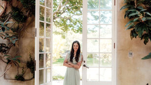 Wallcovering artist Trisha Patel in a pale green dress smiles in a sunlit open doorway, framed by white French doors and leafy vines.