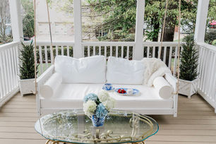 White porch swing with pillows and faux fur blanket, tea and strawberries on blue china, flowers on glass table, leafy yard behind