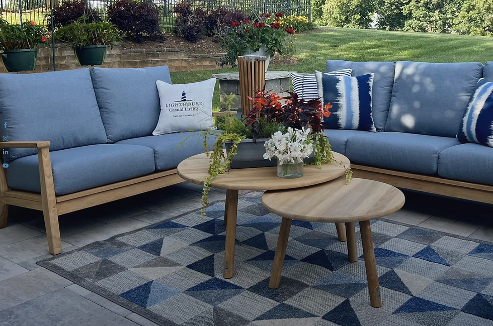 Outdoor patio scene with blue sofas, a wooden table with flowers, a geometric rug, and lush greenery. "Lighthouse Casual Living" pillow visible.