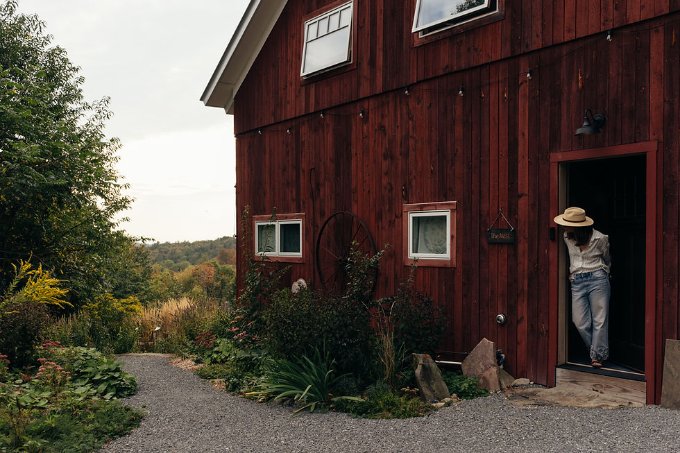 Kate Robinson wearing a straw hat stands in a doorway of a red barn-style house beside a sign reading The Nest, with a garden path and hills.