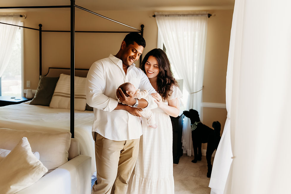 A couple smiles at their baby in a cozy bedroom with white curtains, a bed, and a dog by the window. Bright, warm atmosphere.