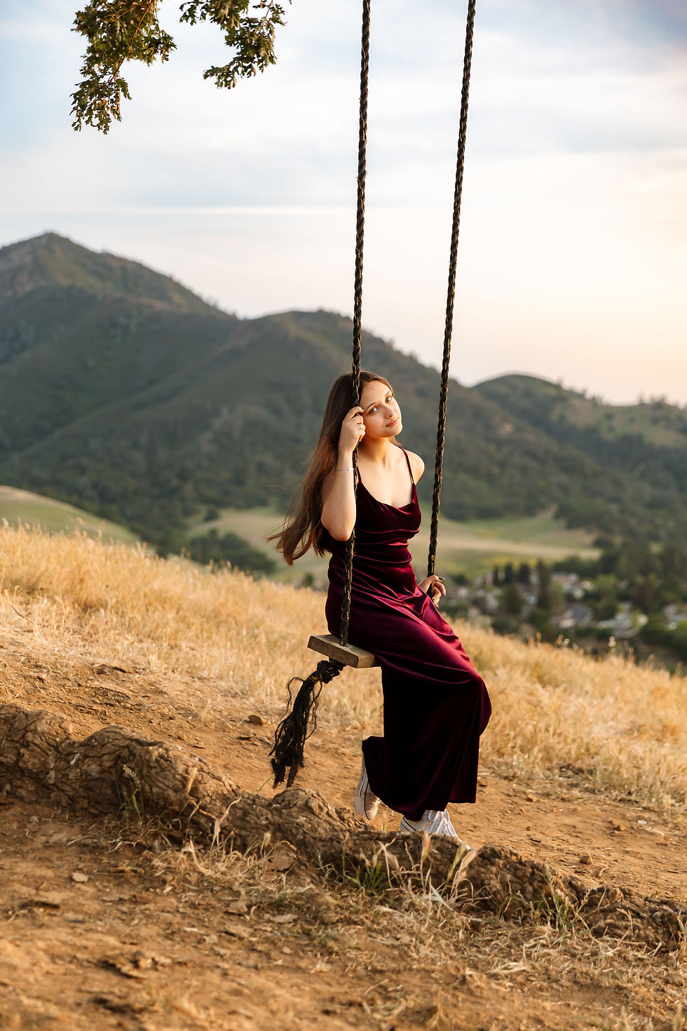 A high school senior woman in a burgundy dress sits on a swing on a grassy hill, with mountains in the background. The mood is reflective and serene.