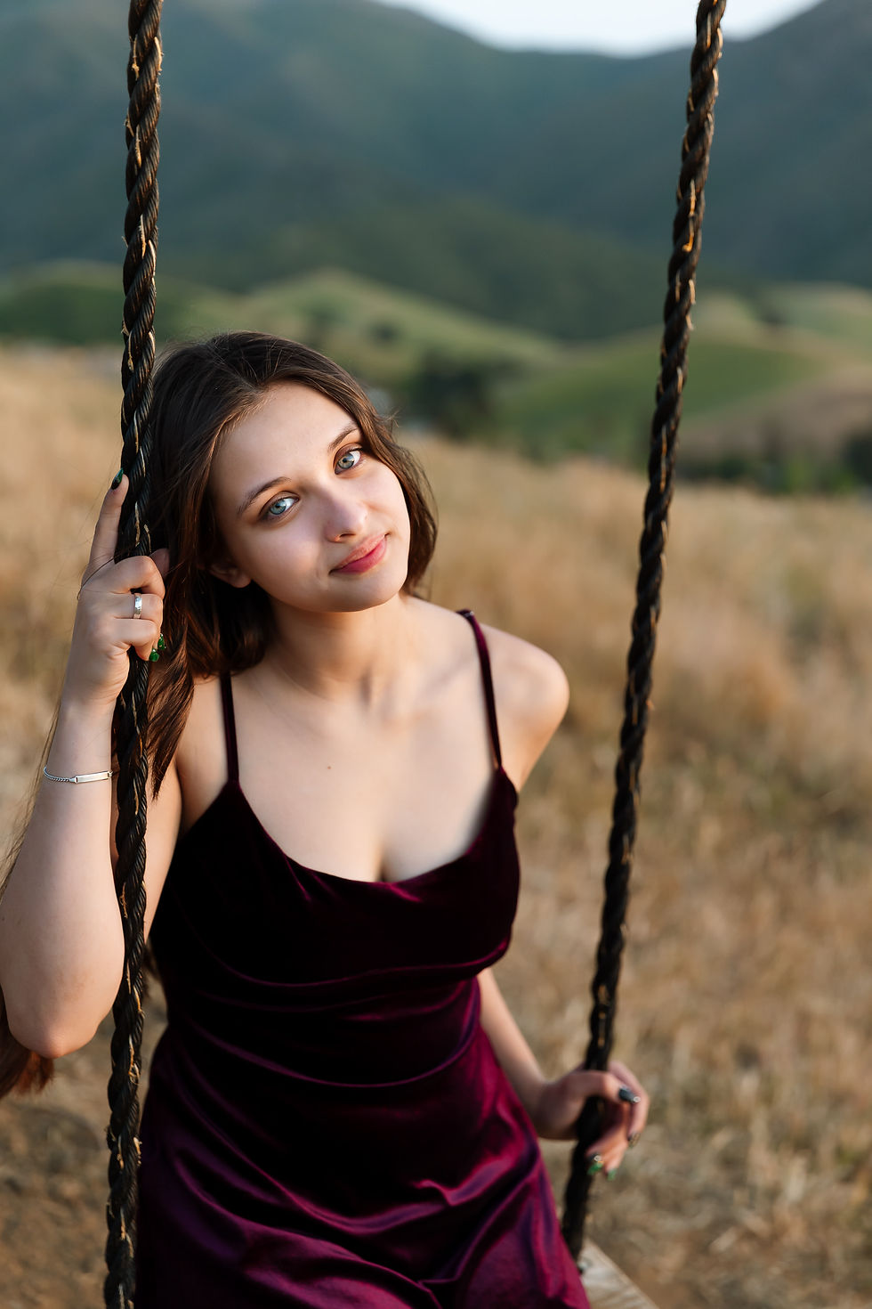 A Clayton Valley Charter High School senior woman in a burgundy dress sits on a swing in a field, with hills in the background. She looks content and relaxed, holding the rope.