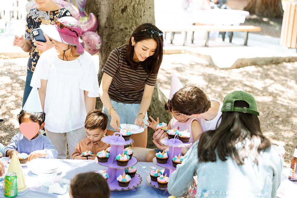 Adults and children gather at an outdoor birthday party with purple cupcake stands. A woman serves cupcakes. Party hats and balloons create a festive mood.