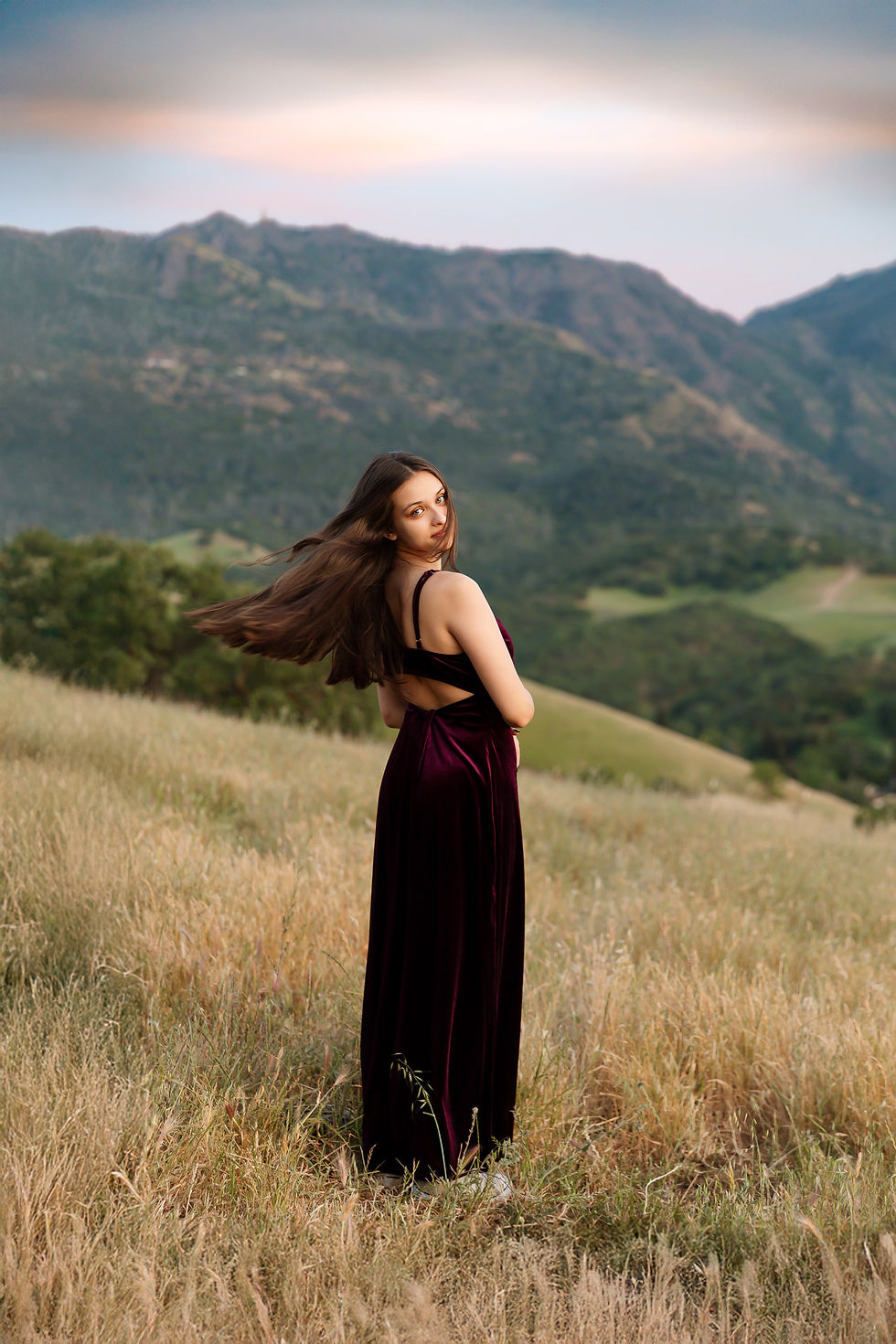 Clayton Valley Charter High School Senior Woman in a burgundy dress stands in a grassy field, looking back with long hair flowing. Mountains and a soft sky are in the background.