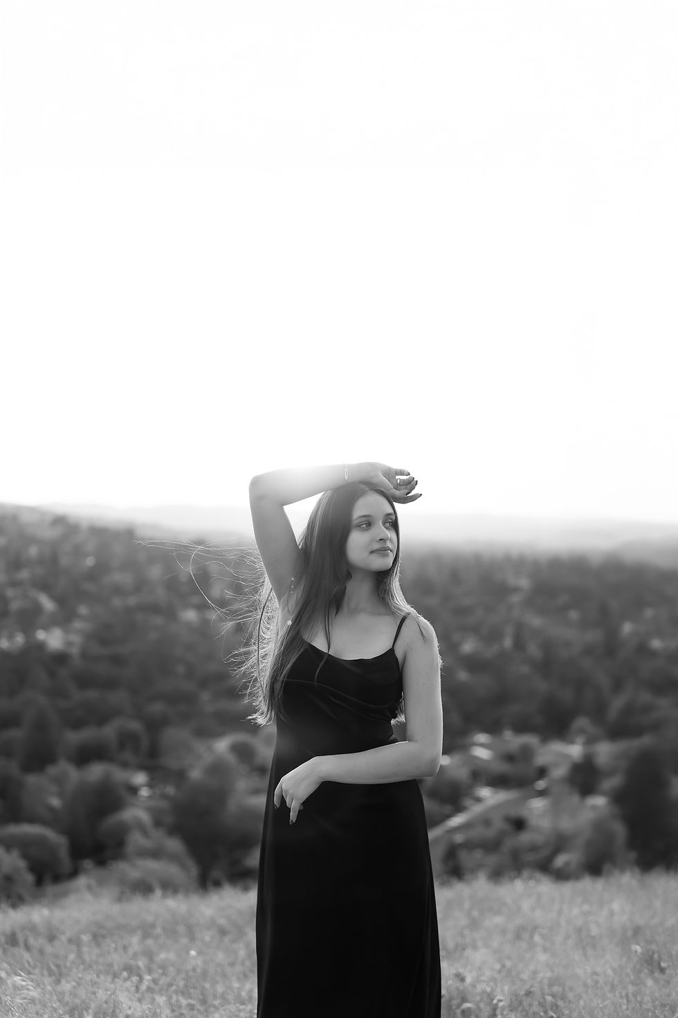 Woman in black dress stands on grassy hill, arm raised, looking contemplative. Background shows blurred landscape under bright sky.