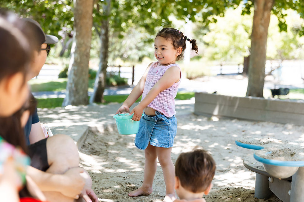A girl in a pink top and denim shorts holds a blue bucket, smiling in a sunny park playground. Other children and trees are visible.
