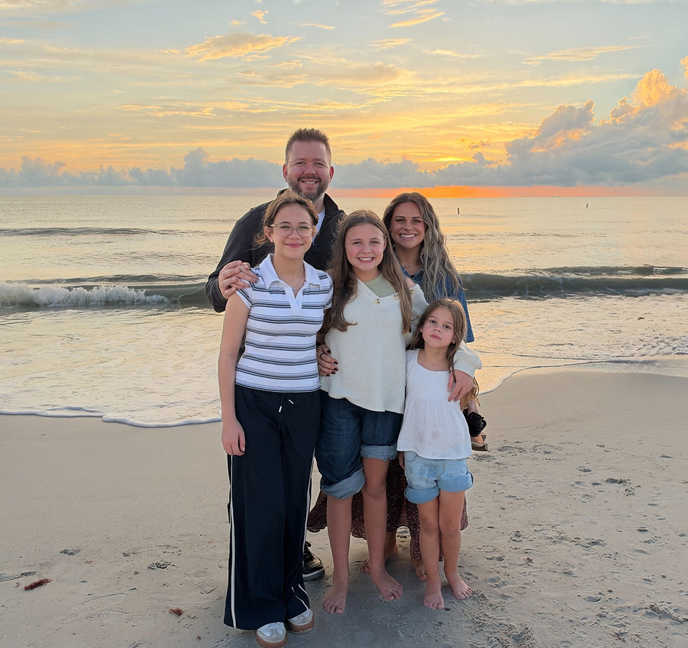 A smiling family of five standing together on the beach at sunset, with gentle waves and a golden sky behind them.